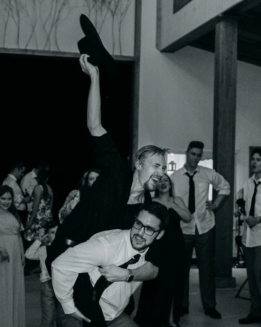 People at a lively indoor gathering, with two men in the foreground, one holding the other in a piggyback position while both laugh. Other guests are seen in the background, some standing and talking, some smiling and watching.
