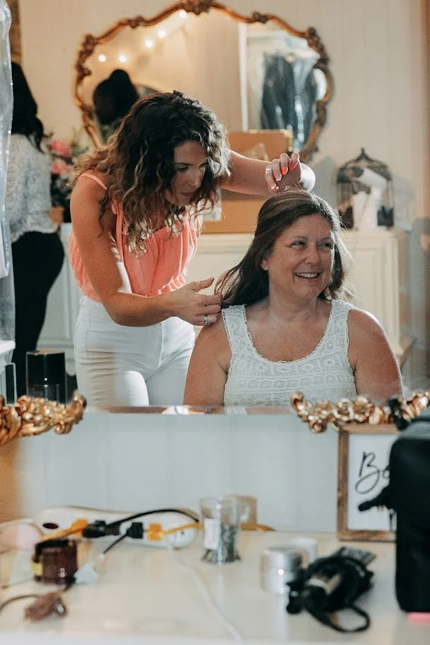 A woman sitting at a vanity mirror being styled by another woman, both smiling, in a warmly decorated room.