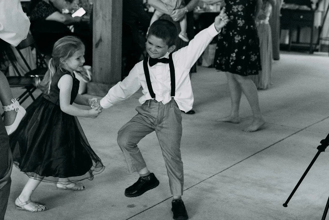 A young boy and girl dancing together at a social event. The boy is wearing a white shirt, bow tie, suspenders, and pants, while the girl is dressed in a dark dress with a ribbon in her hair. They are holding hands and appear joyful, with other peopl