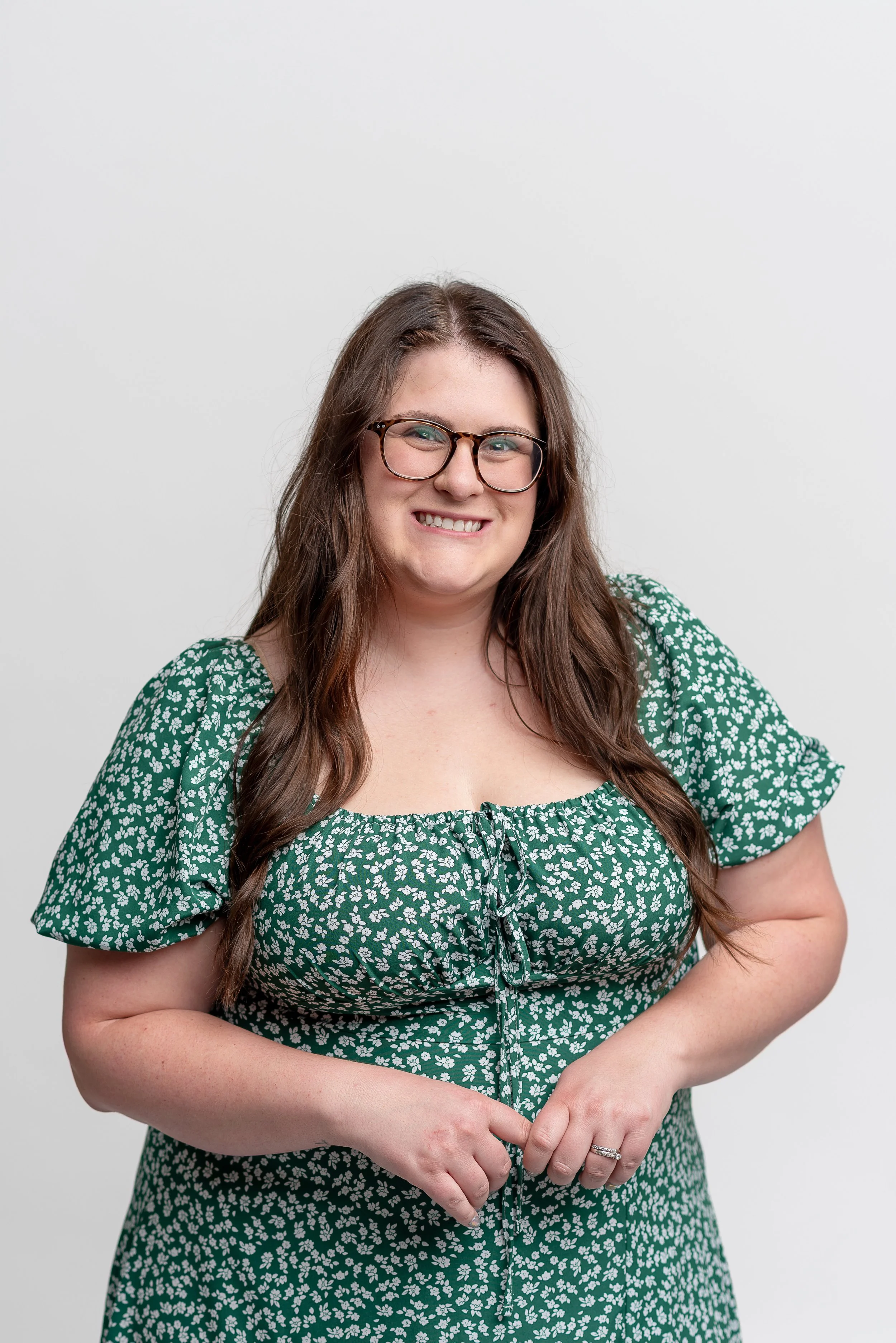 A woman with long brown hair wearing glasses and a green floral dress, smiling and standing against a white background.