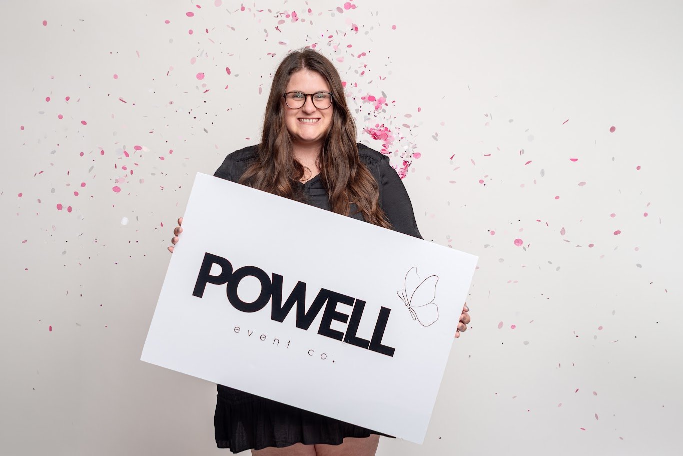 A woman with long brown hair and glasses smiling while holding a large white sign with the words "POWELL event co." and a butterfly drawing, with pink and gray confetti in the background.