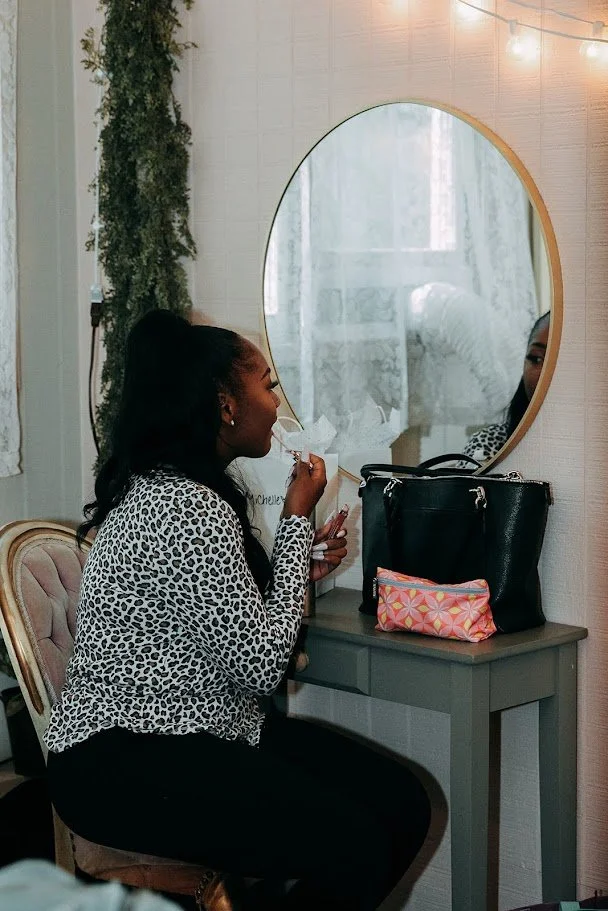 A woman with long dark hair in a top knot, wearing a leopard print top, sitting on a pink upholstered chair, looking into a round mirror while applying lipstick. There is a black handbag and pink patterned pouch on the gray table beside her. The wall