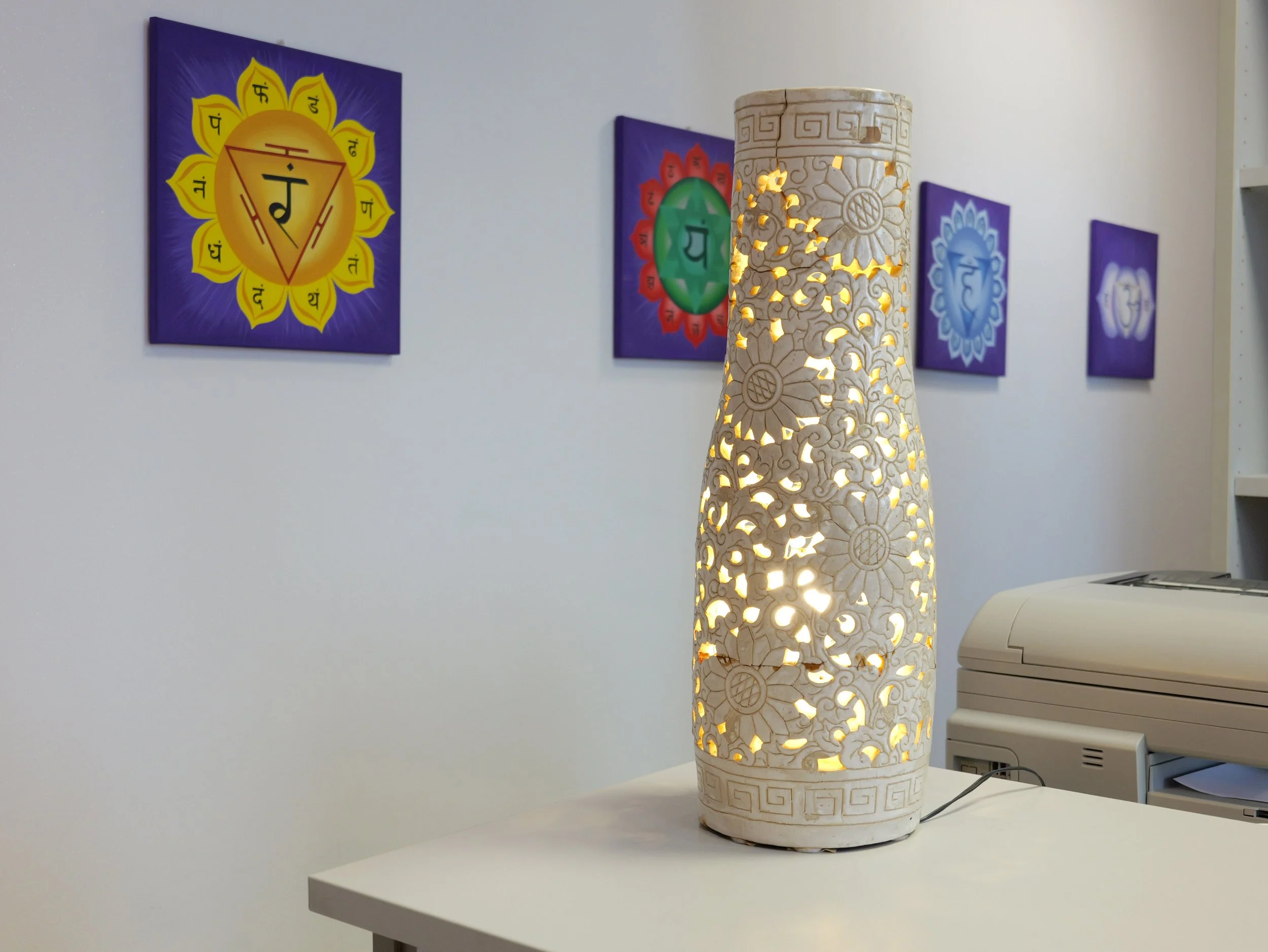 Lampada decorativa bianca con disegni traforati emette luce calda. Sul muro, quattro quadri con simboli e pronunce di origine indiana o buddista. Studio Olistico Geode Modena