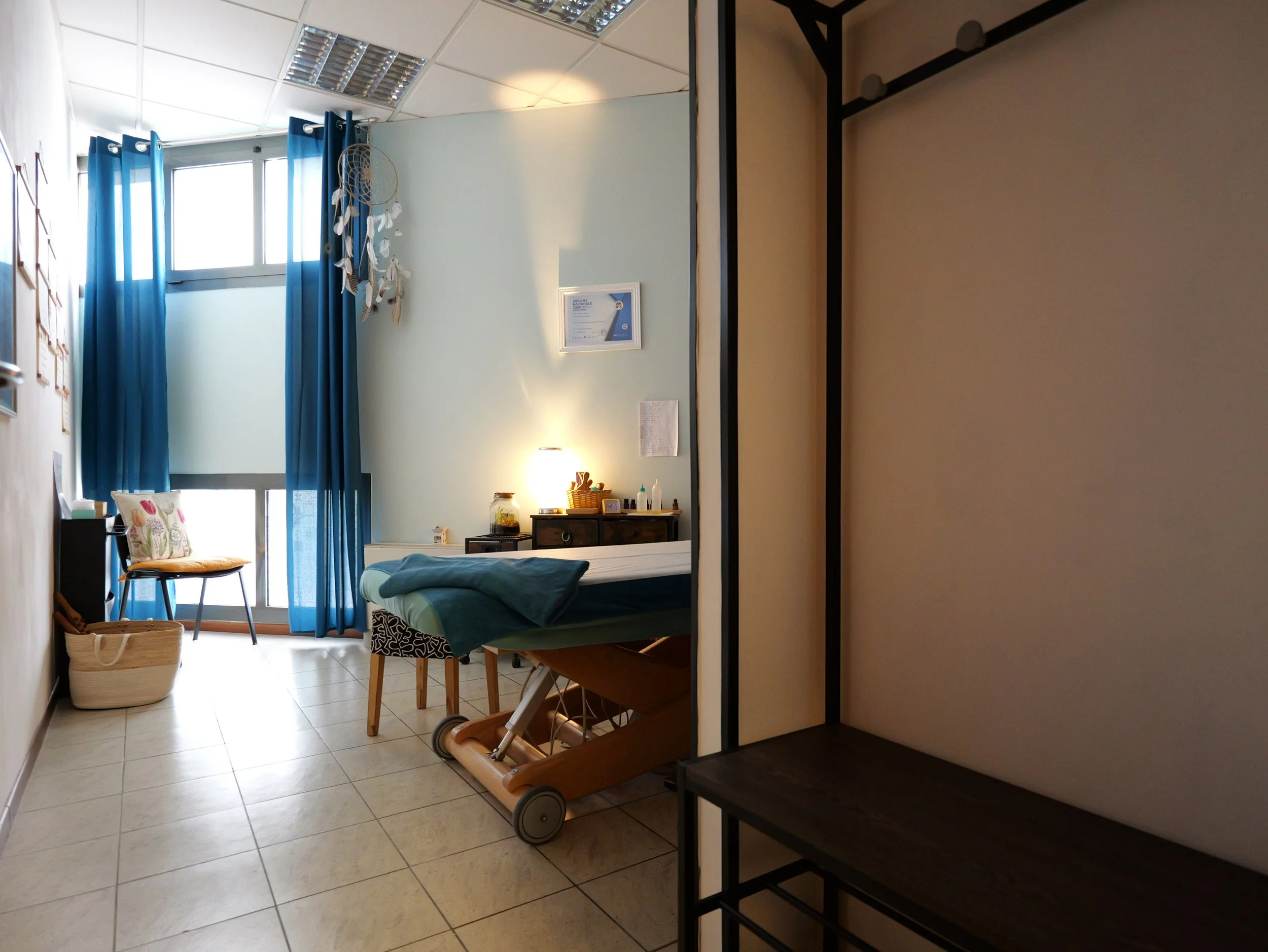 Interno di una stanza con pareti di colore celeste, con una finestra con tende blu, un letto, una sedia con cuscino decorato, una consolle con oggetti e una lampada. Studio Olistico Geode Modena
