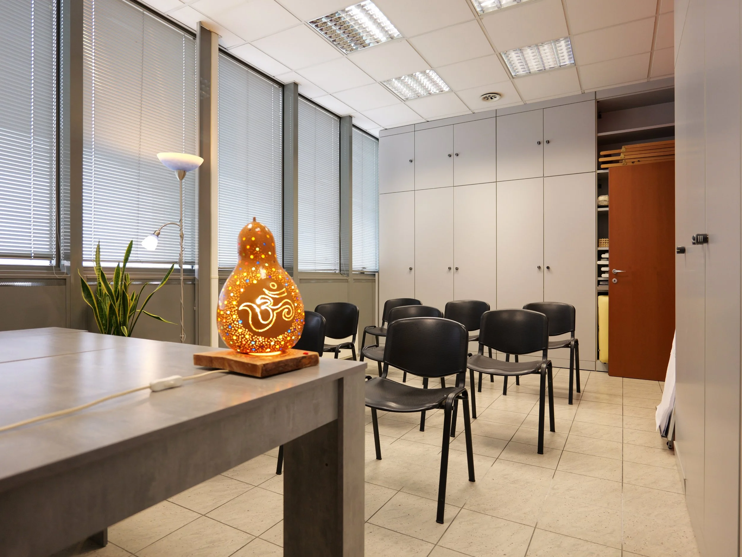 Sala riunioni con sedie nere, armadi bianchi e finestre con tende orizzontali. Decorazioni tra cui una lanterna decorativa luminosa con simbolo OM e una pianta. Illuminazione con luci al neon. Studio Olistico Geode Modena