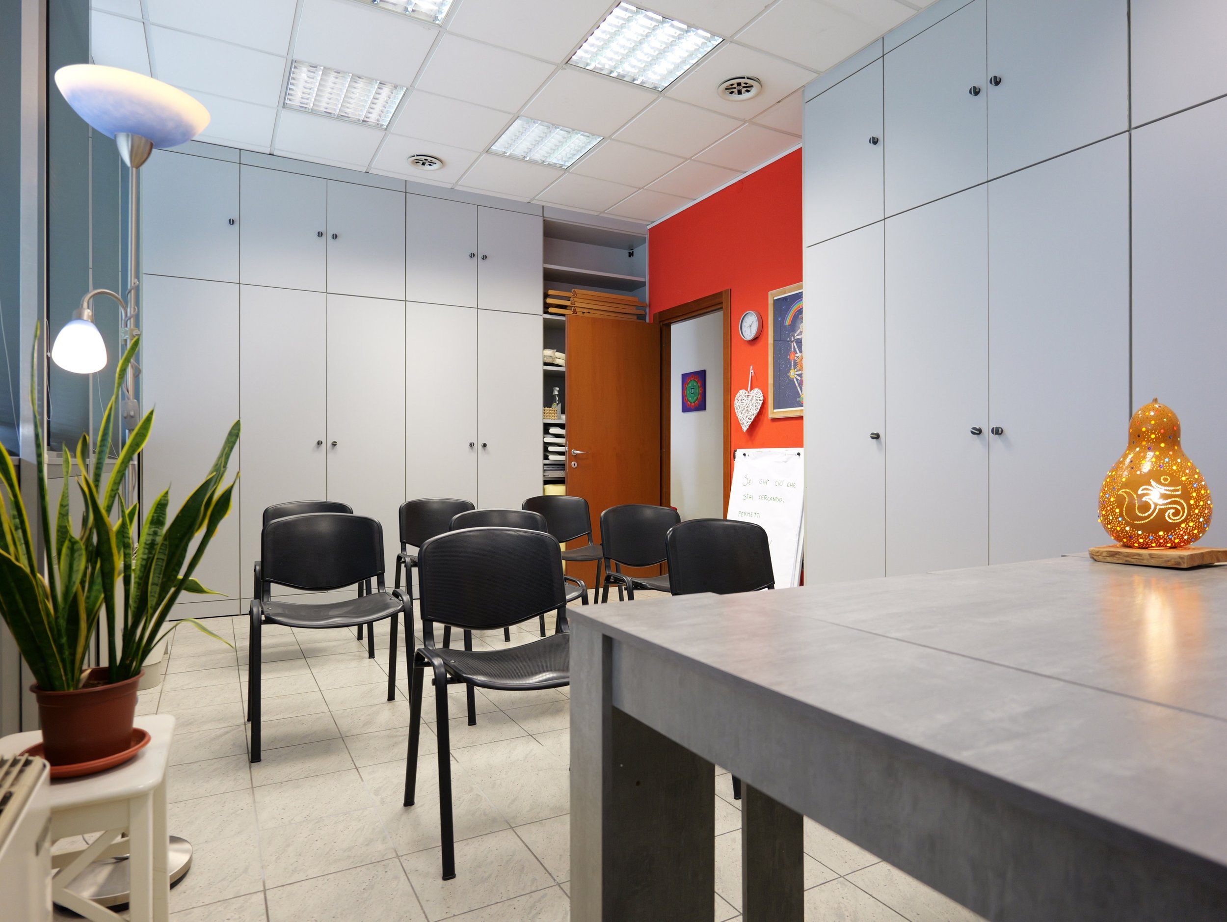 Sala di riunione con pareti bianche, soffitto con luci fluorescenti, sedie nere disposte in cerchio, piante verdi, decorazioni colorate e lampada ornamentale arancione. Studio Olistico Geode Modena
