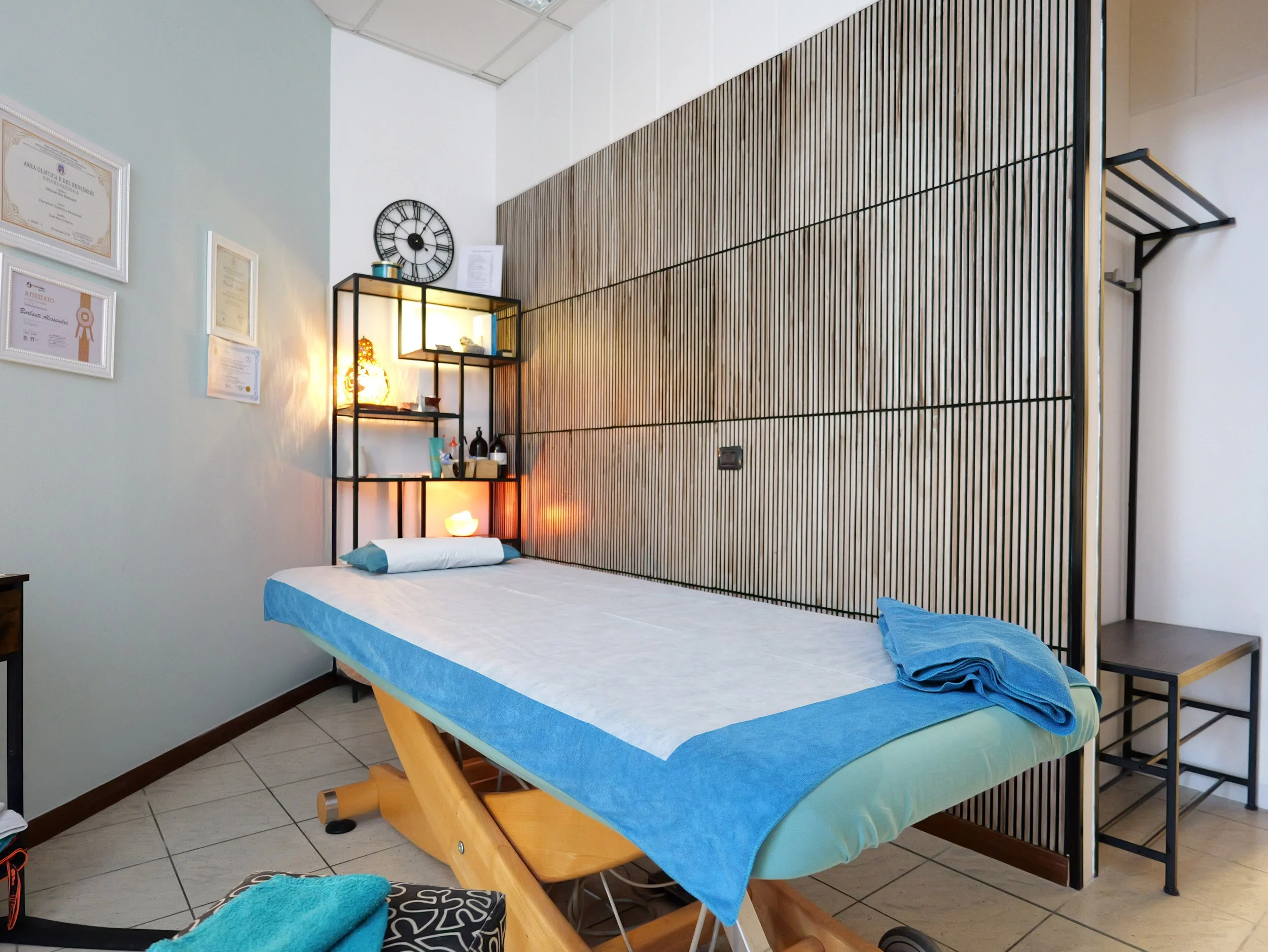 Letto da massaggi con lenzuola bianche e coperte blu, in una stanza con pareti bianche e una parete di rivestimento in legno, decorata con quadri e un orologio. Studio Olistico Geode Modena