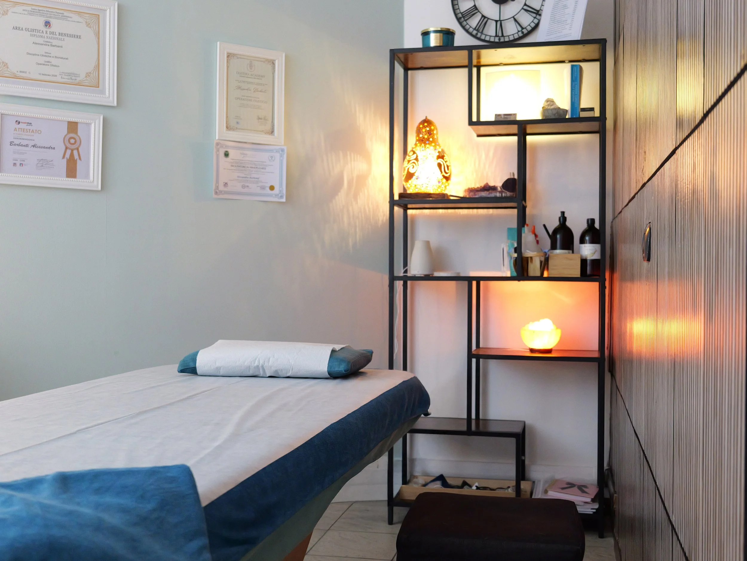 Stanza con un letto con lenzuola bianche e cuscino grigio, scaffale nero con lampada che emette luce calda, certificate appese al muro, parete di legno e parete di colore chiaro. Studio Olistico Geode Modena
