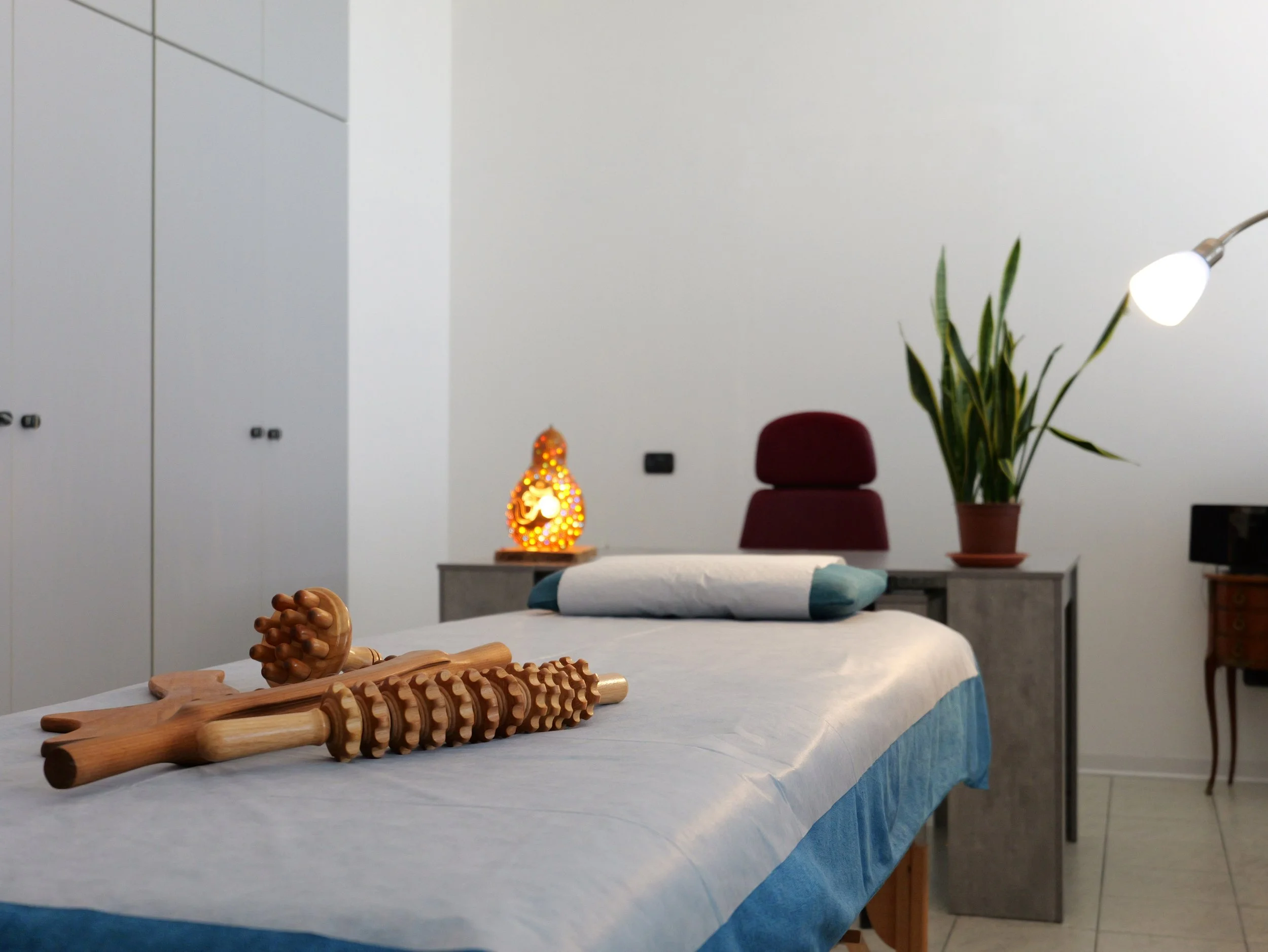 Tavolo coperto con una tovaglia bianca, decorazioni in legno e una ciotola con un fiore artificiale. Sul tavolo ci sono strumenti di massaggi in legno. Studio Olistico Geode Modena