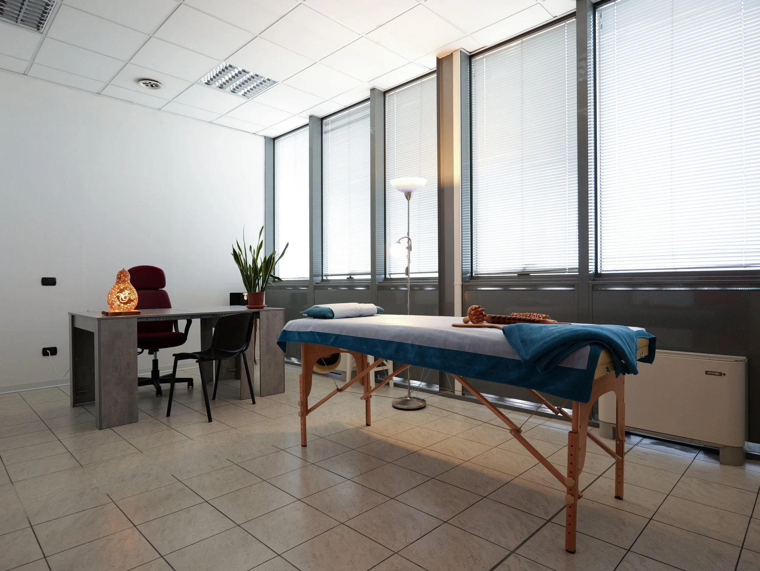 Stanza di terapia oolistica, un lettmassaggi blu, lampada da terra, parete con grandi finestre e tapparelle in posizione semi chiusa, piante e decorazioni sul tavolo. Studio Olistico Geode Modena