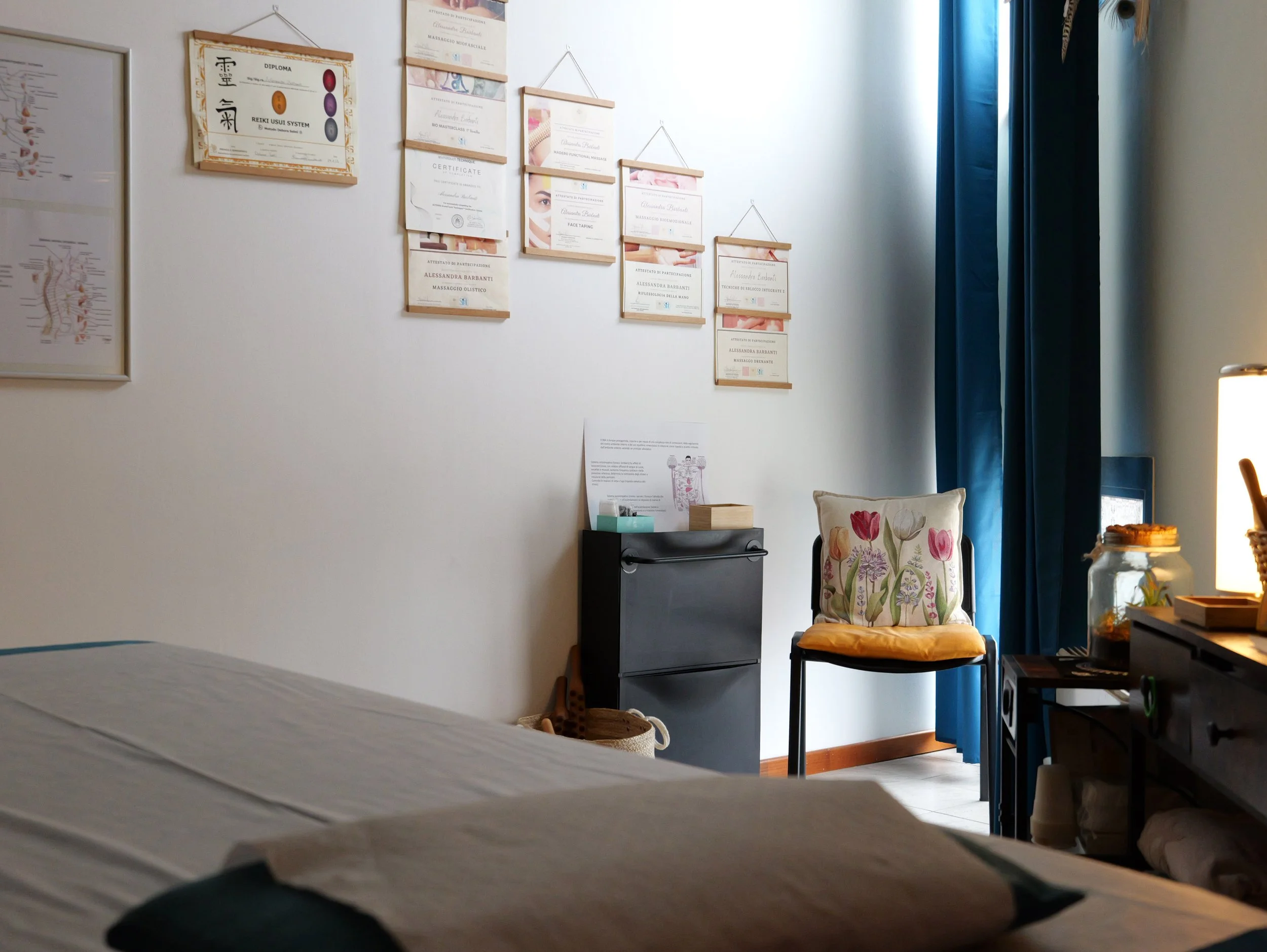 Una parete con diverse diplomi e attestati incorniciati, una sedia con cuscino decorato con fiori, una finestra con tende blu, un tavolo con alcuni oggetti e una lampada. La stanza è uno studio di massaggi. Studio Olistico Geode Modena