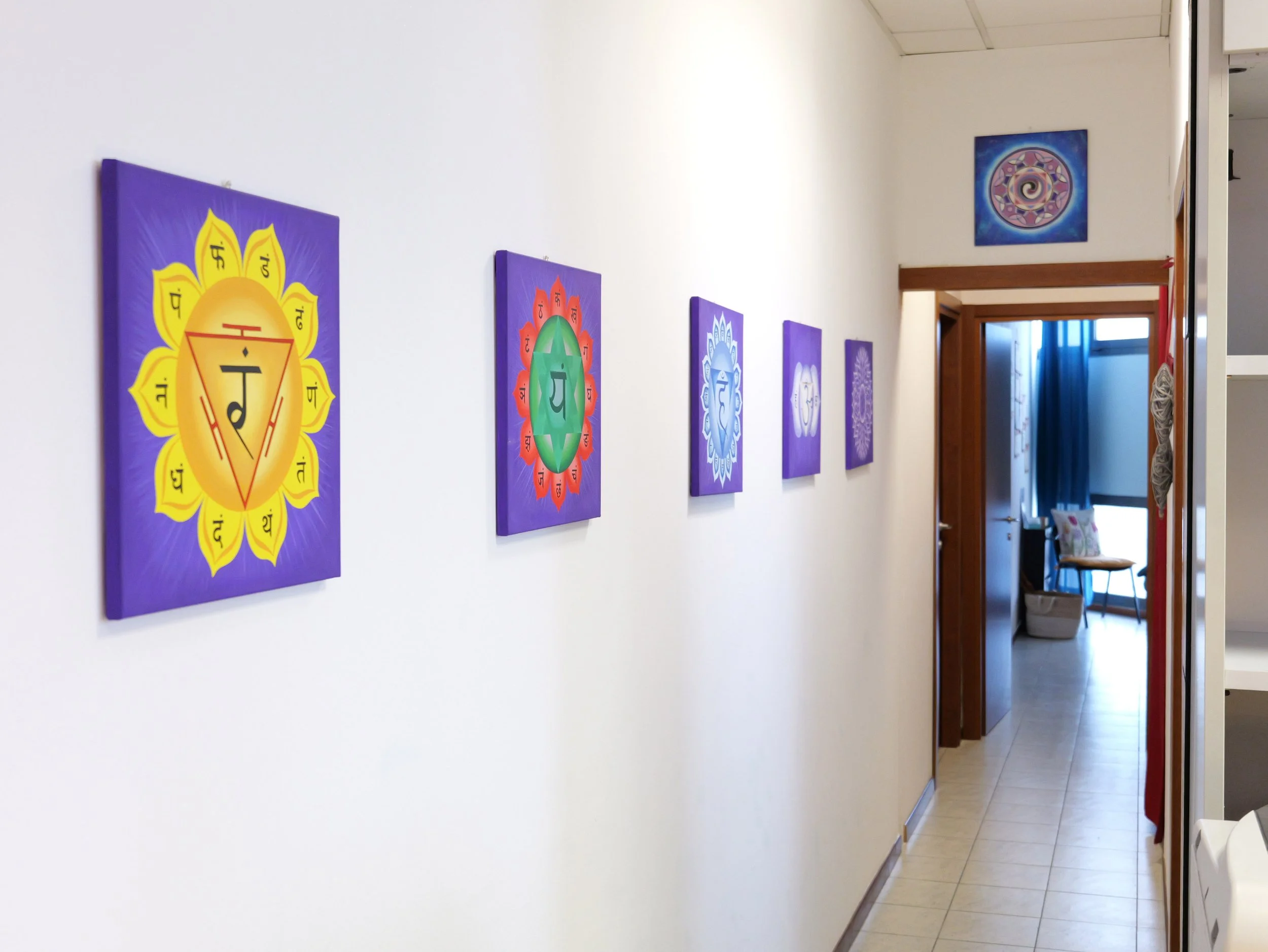 Una parete bianca con dipinti colorati raffiguranti simboli di chakra lungo il corridoio di un interno, con una porta e una finestra alla fine del corridoio. Studio Olistico Geode Modena