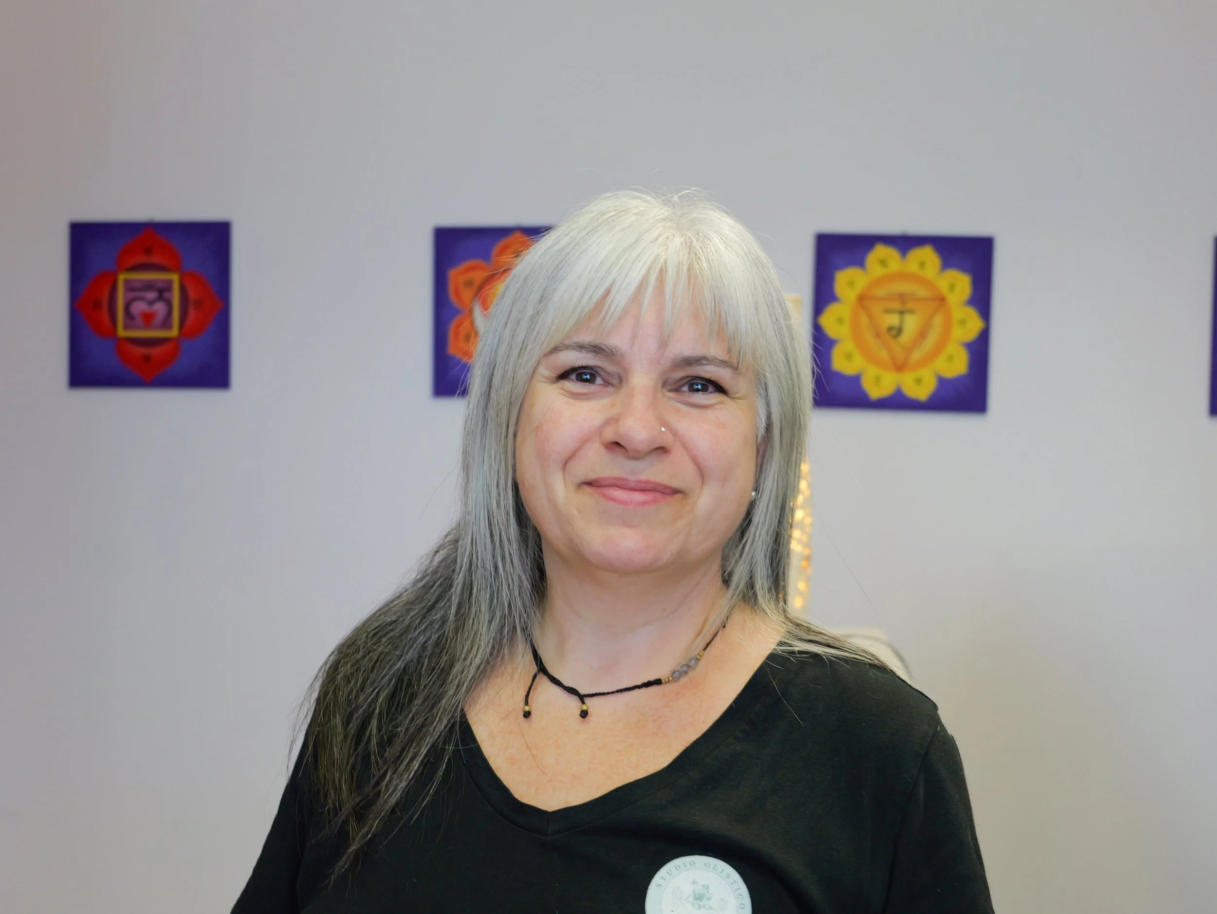 Una donna con capelli grigi, indossa una maglietta nera con un logo sul petto e una collana. Sullo sfondo, tre quadri colorati con simboli spirituali. Alessandra Barbanti Studio Olistico Geode Modena