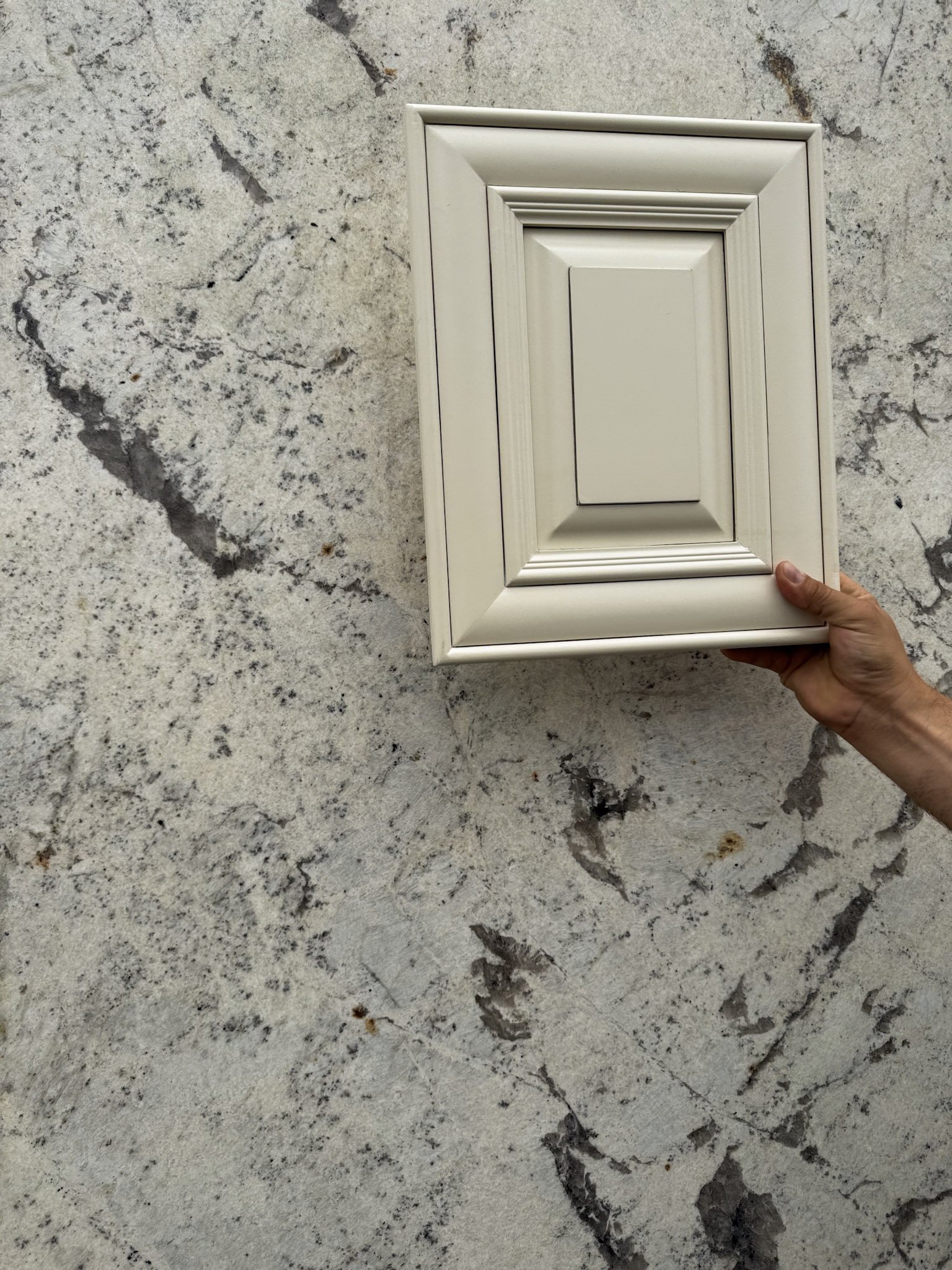 A person holding a cream cabinet sample over a granite stone countertop.