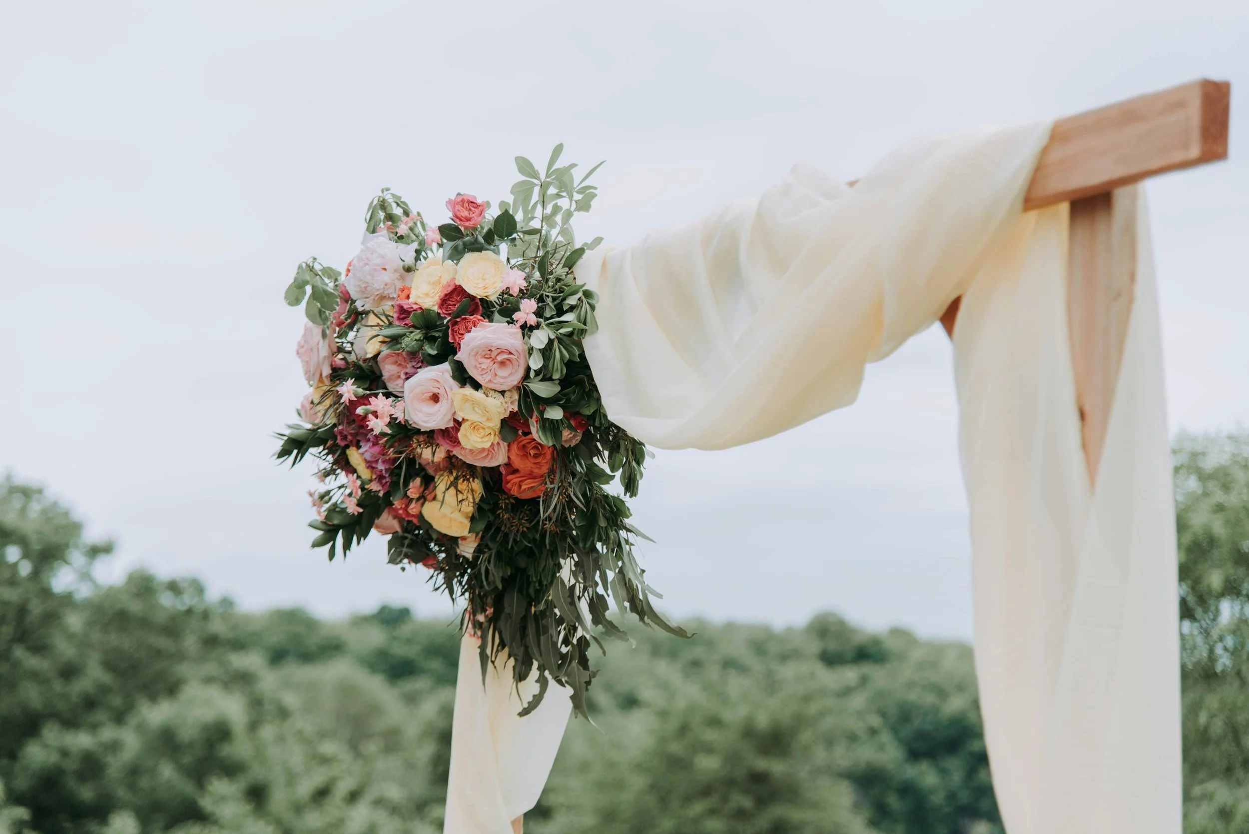 A bouquet of pink, cream, and orange flowers attached to a wooden stand draped with cream fabric outdoors with green trees in the background.