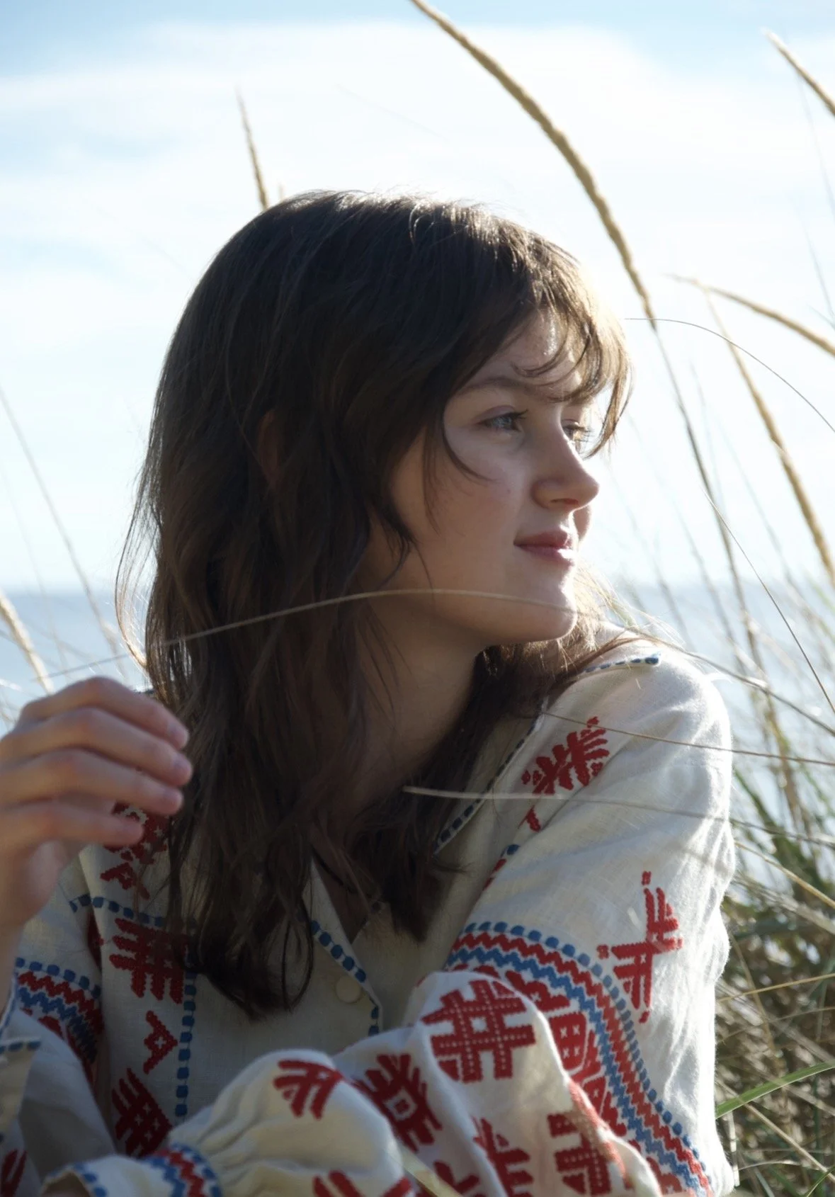 A young woman with wavy brown hair and light skin sitting outdoors in a grassy area, gazing thoughtfully into the distance, wearing a beige shirt with red and blue embroidery.