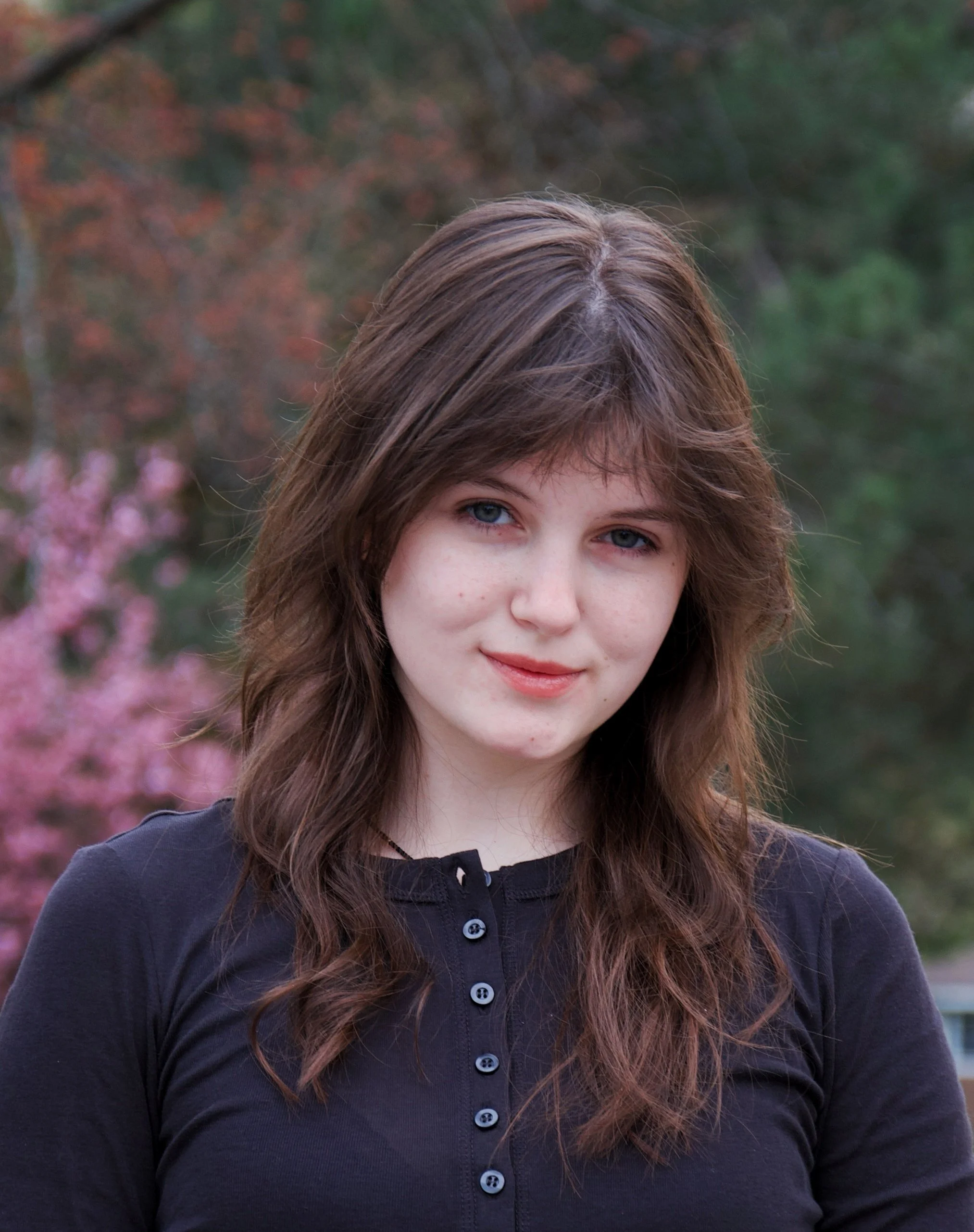 Close-up of a young woman with light skin and long, wavy brown hair standing outdoors with pink flowering trees and green shrubbery in the background.