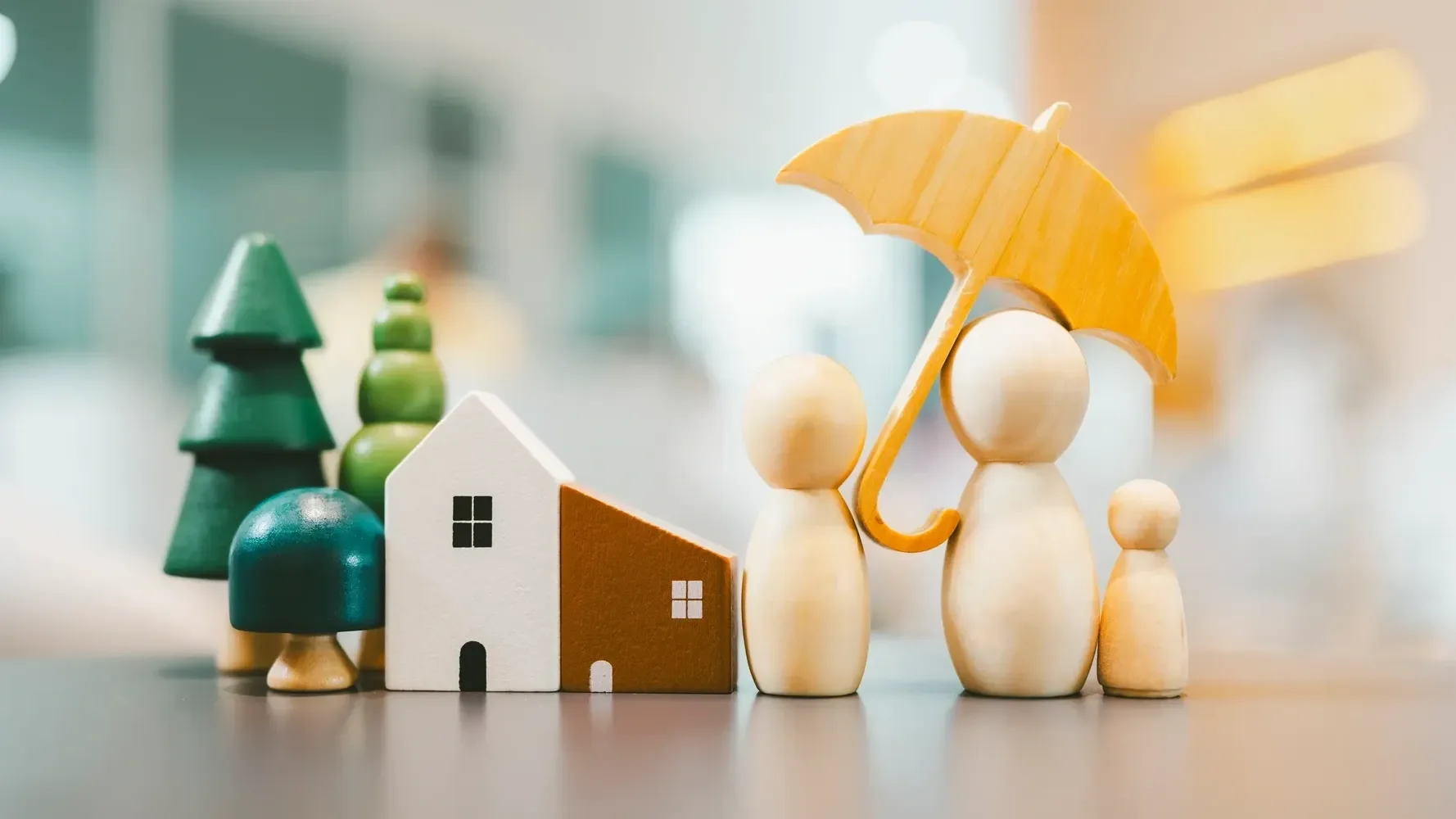 Wooden toy figures, including a house, trees, and people holding a yellow umbrella, on a blurred indoor background.