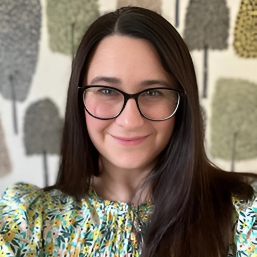 A woman with long dark hair and glasses smiling at the camera, wearing a floral-patterned top, with a wall decorated with tree-shaped art in the background.