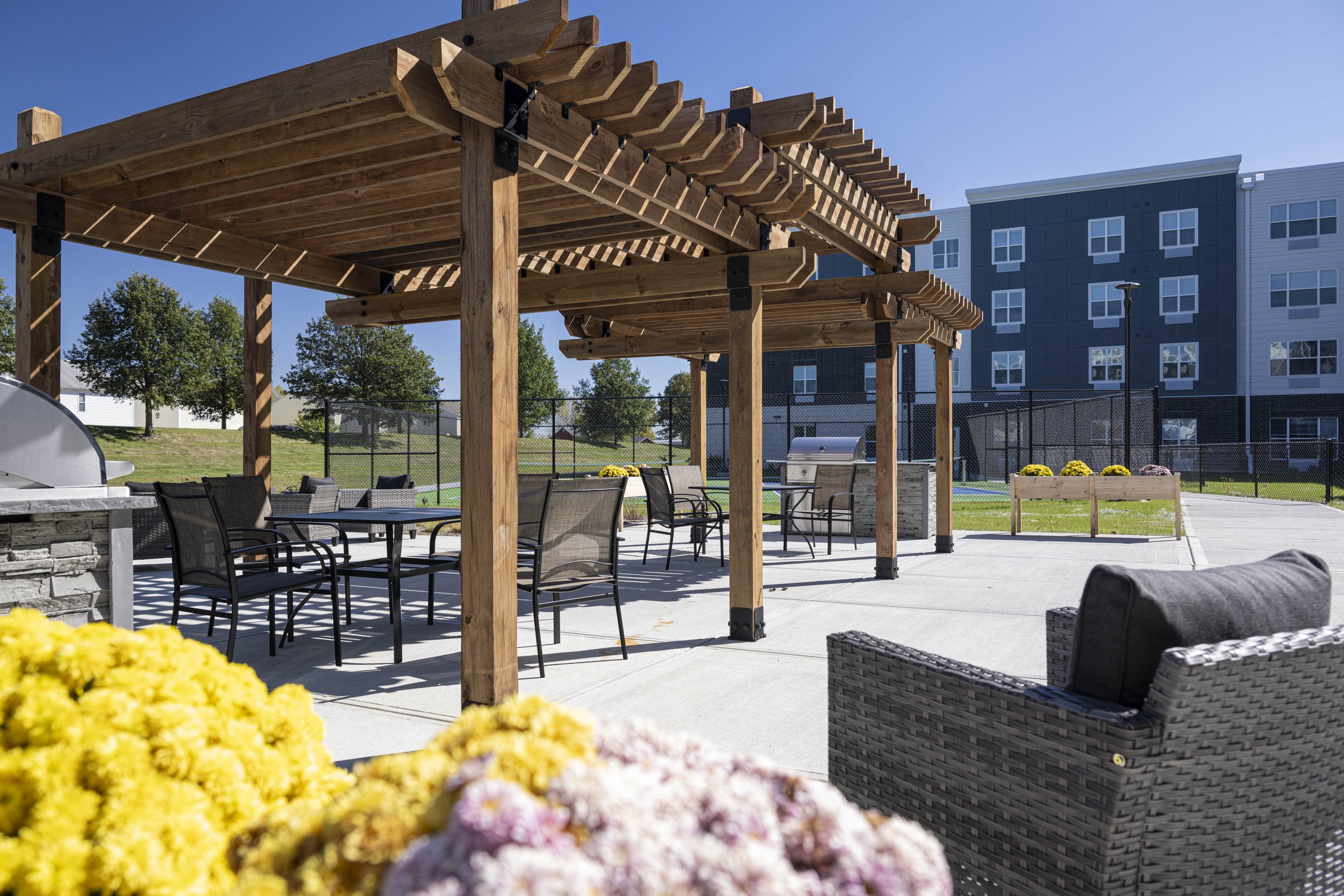An outdoor patio area with wooden pergolas covering a table and chairs, a built-in grill, and colorful flower planters, with a modern apartment building and a chain-link fence in the background under a clear blue sky.