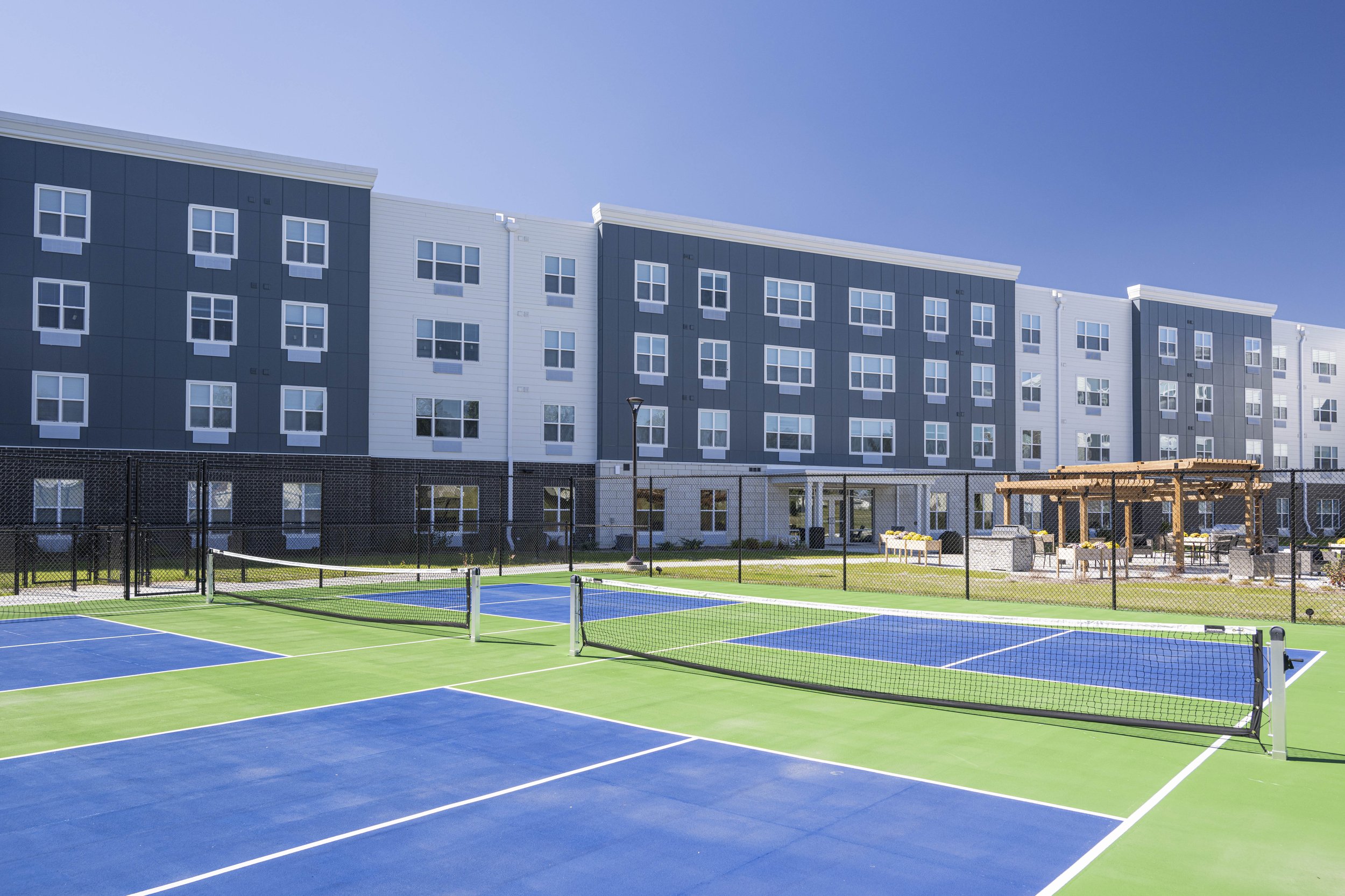 A view of a modern apartment complex with blue and green outdoor tennis courts, a pergola, and outdoor seating under clear blue sky.