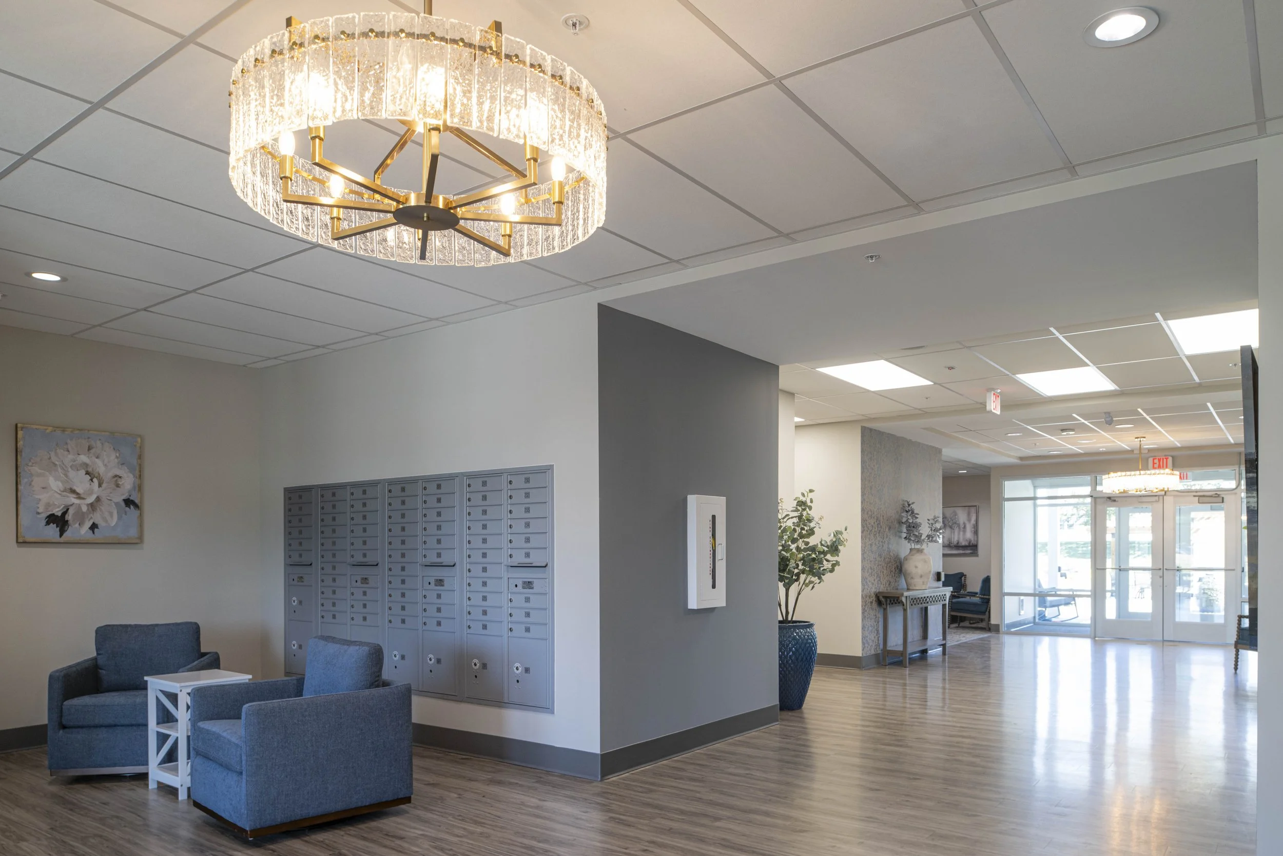 Lobby area with white walls, wooden flooring, a modern chandelier, two blue armchairs with a small white table, decorative art on the wall, large potted plants, storage lockers, and glass doors at the entrance.
