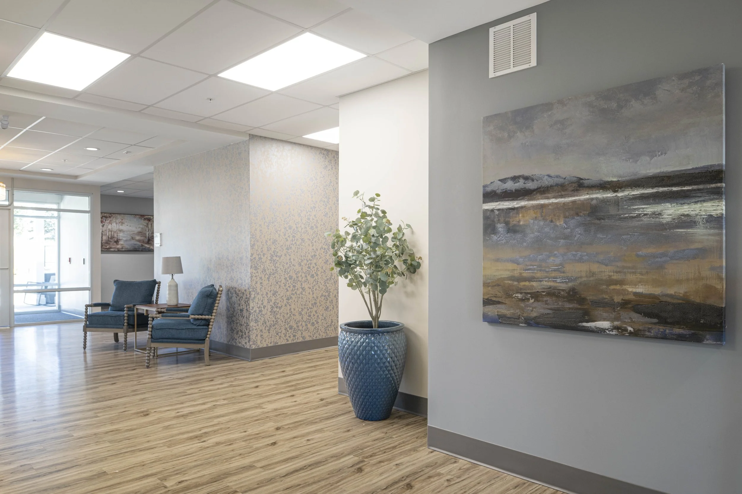 Modern waiting area with two blue armchairs with patterned upholstery, a small wooden table, a white table lamp, a potted plant in a large blue ceramic pot, and abstract landscape artwork on the wall. The space has light-colored wooden flooring and a drop ceiling with square fluorescent lights.