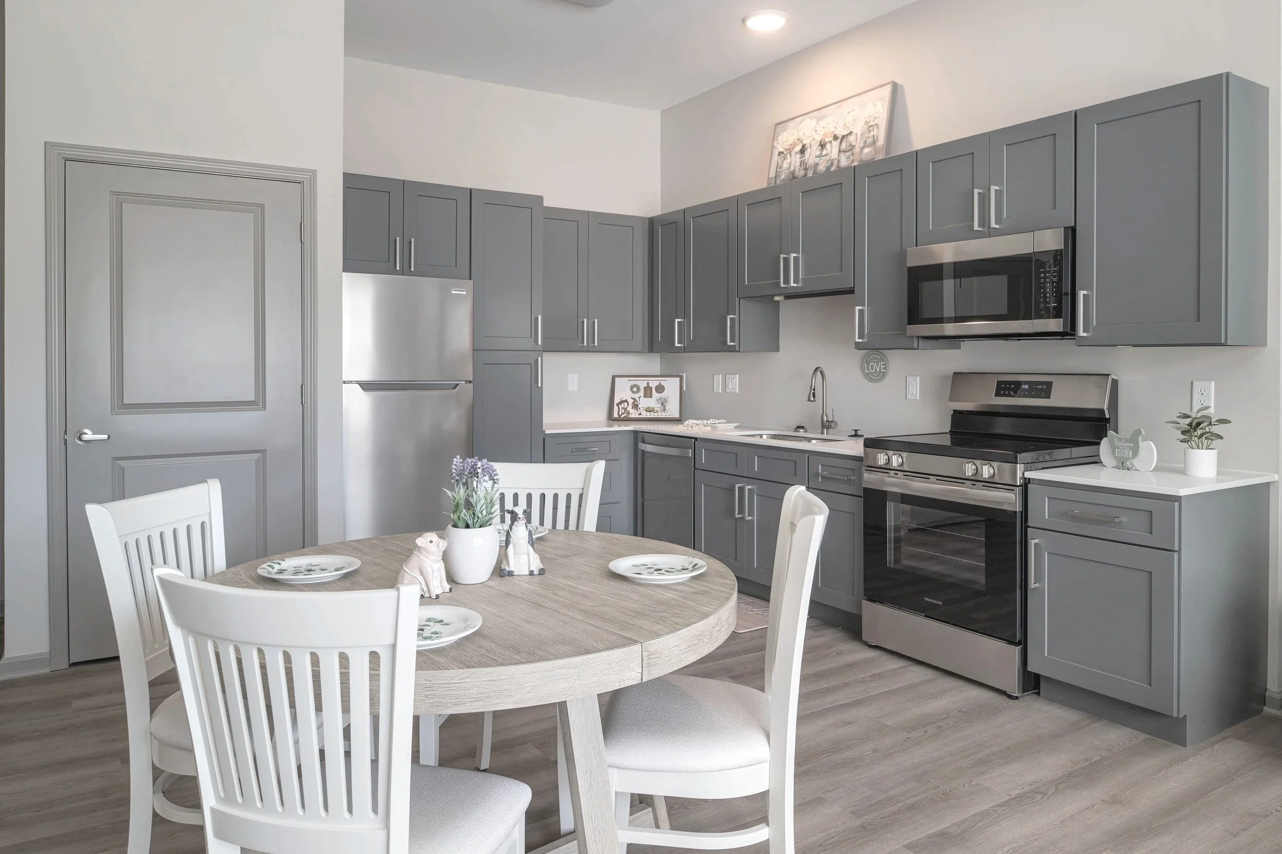 A modern kitchen with grey cabinets, stainless steel appliances, a round dining table with white chairs, and decorative items including a vase with purple flowers and animal figurines.