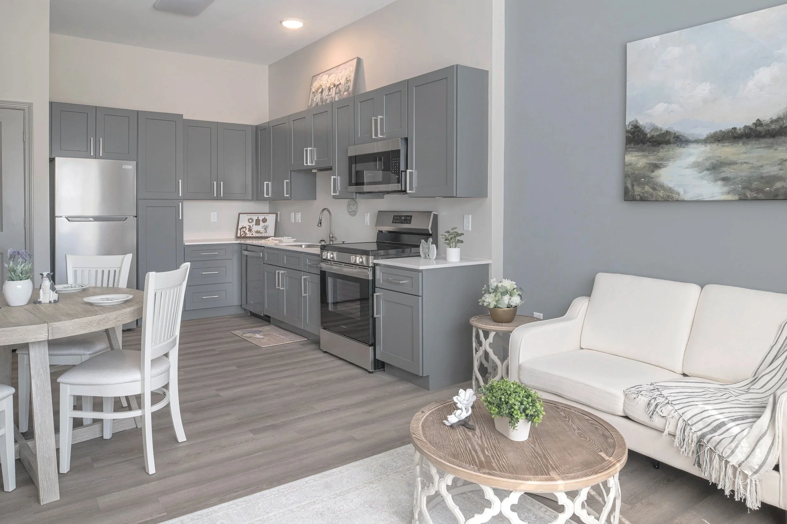 Modern kitchen and living area with gray cabinets, stainless steel appliances, a small round wooden coffee table with white ornate legs, a white sofa with striped pillows, a dining table with white chairs, and wall art featuring a landscape and floral painting.