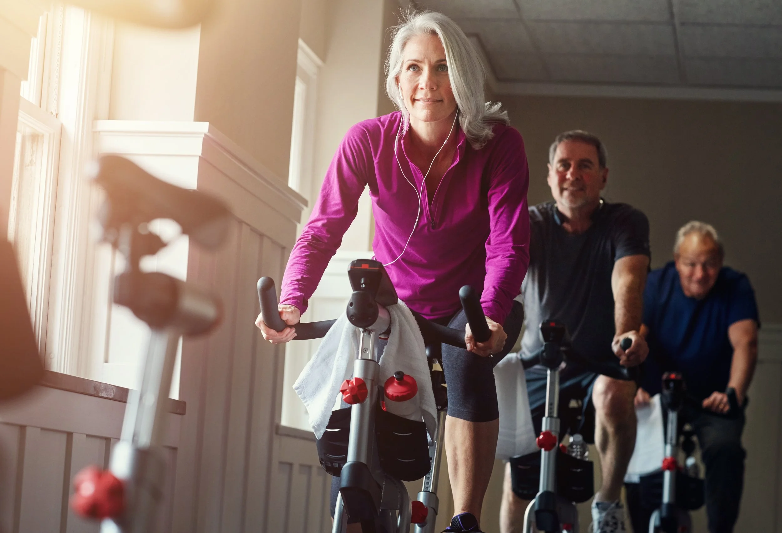 A group of older adults participating in a spinning exercise class indoors, with one woman in a pink top leading the activity and other participants following behind.