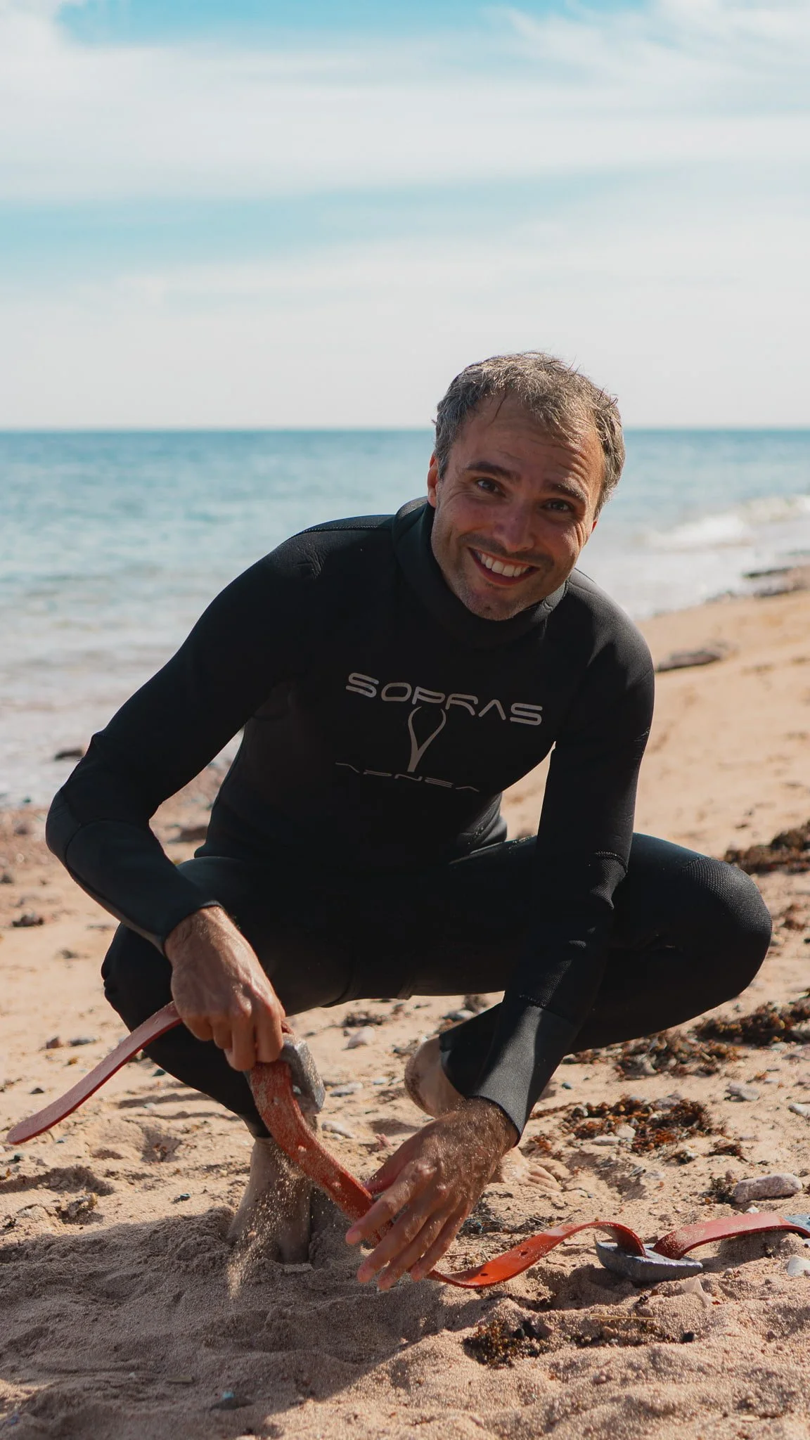 A man in a wetsuit crouches on the sandy beach near the ocean, smiling at the camera while assembling his weight belt. The sky is partly cloudy.