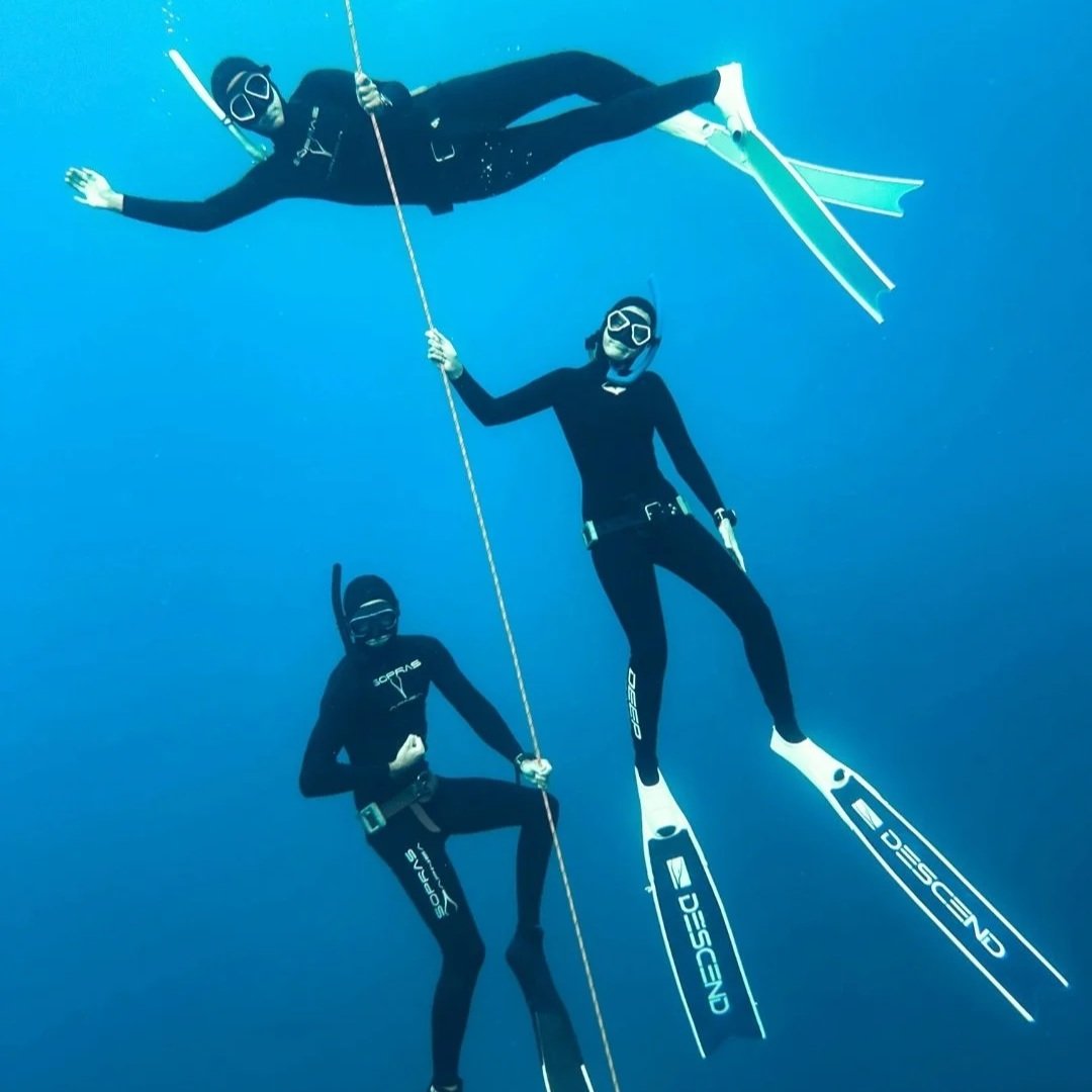 Three scuba divers wearing black wetsuits, masks, and fins underwater, holding onto a rope.