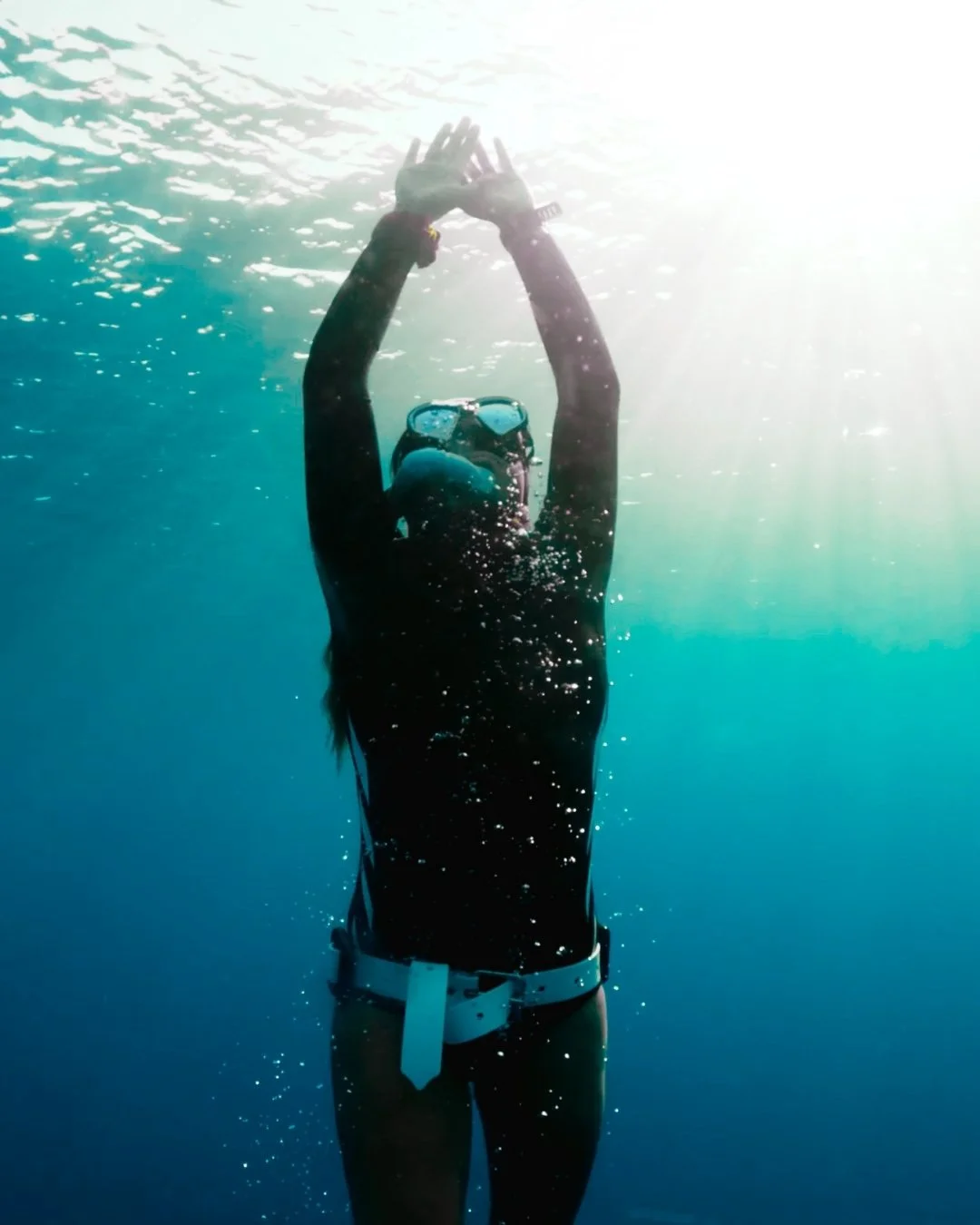 A person freediving underwater wearing a black wetsuit, snorkeling mask, and white weight belt, reaching upward with hands clasped together, illuminated by sunlight from above.