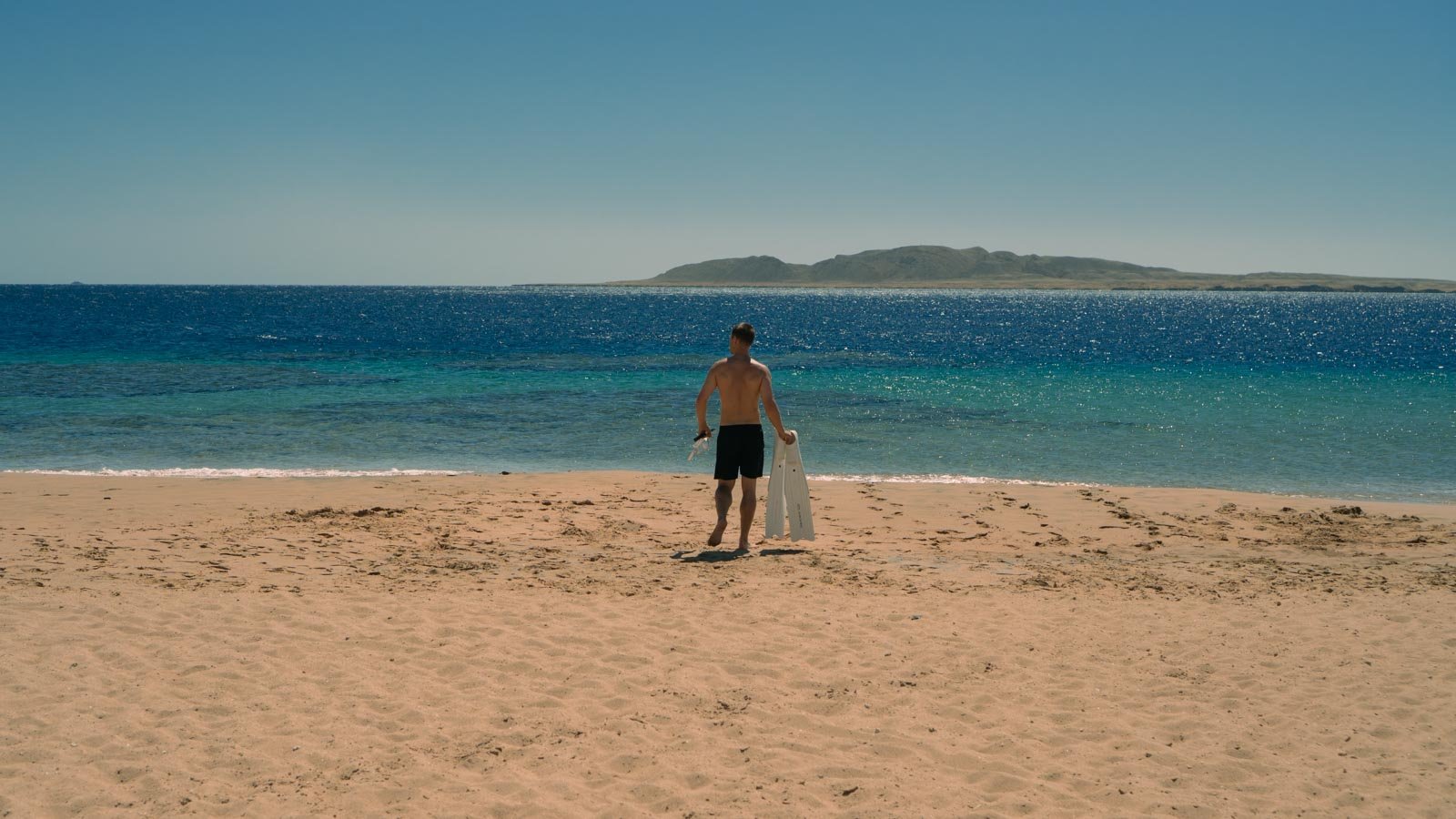 man walking into clear blue sea from shore holding freediving fins