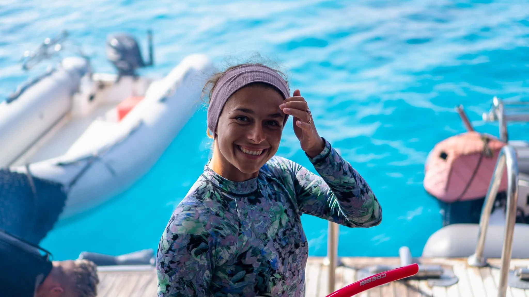 Smiling woman with a headband and floral shirt on a boat by the water, holding a red skate, with a pink bag and some boat equipment nearby.