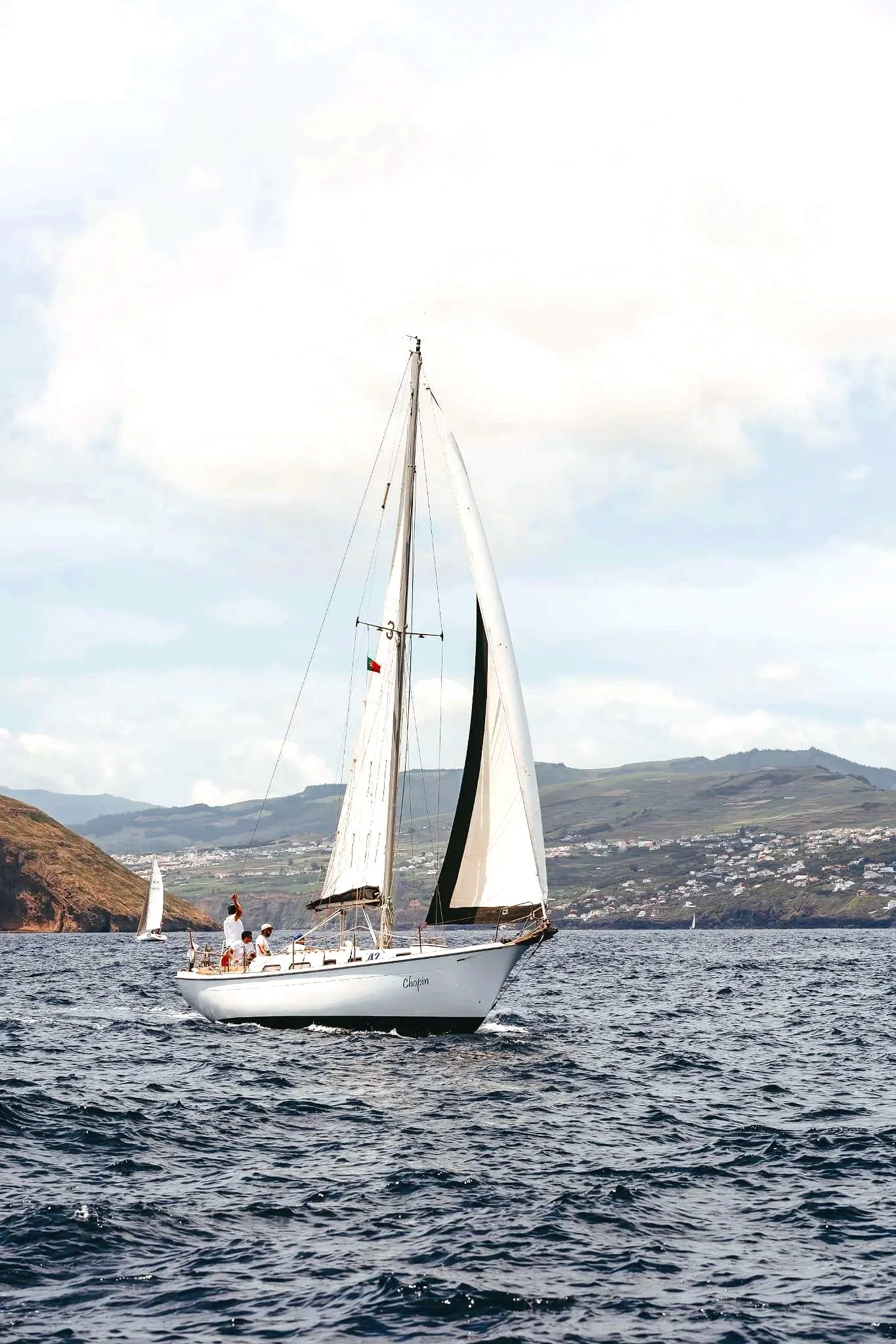 A white sailboat named with four people onboard sailing on the water, with hills of the Azores islands in the background under a partly cloudy sky.