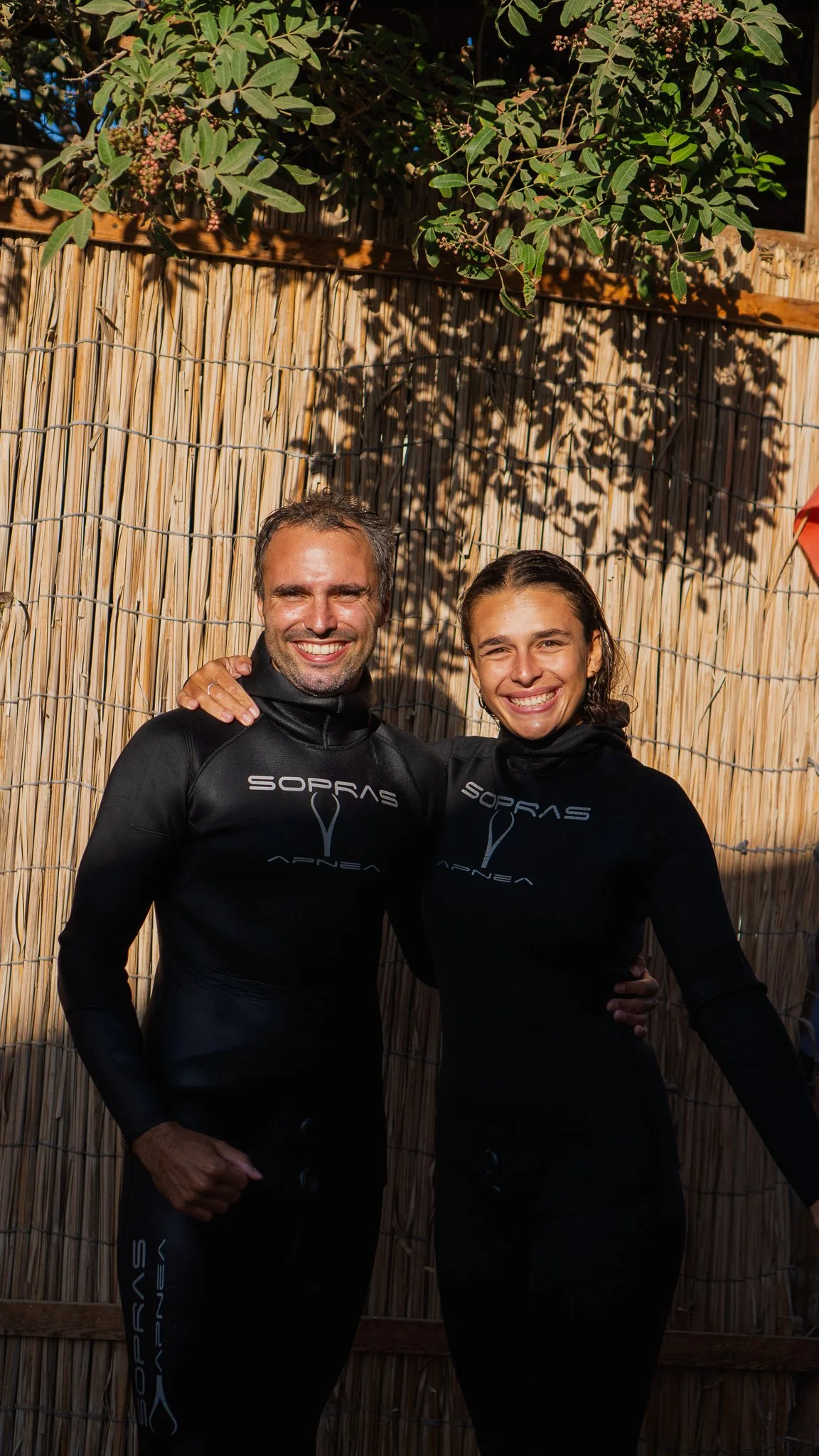 A smiling man and woman dressed in black wetsuits, standing close together in front of a bamboo fence with leaves and pink flowers overhead.