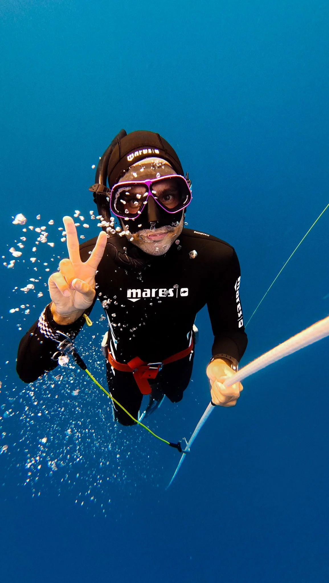 Male freediver underwater near the surface, making a peace sign with his hand, wearing a black wetsuit, purple snorkel mask, and holding a snorkel.