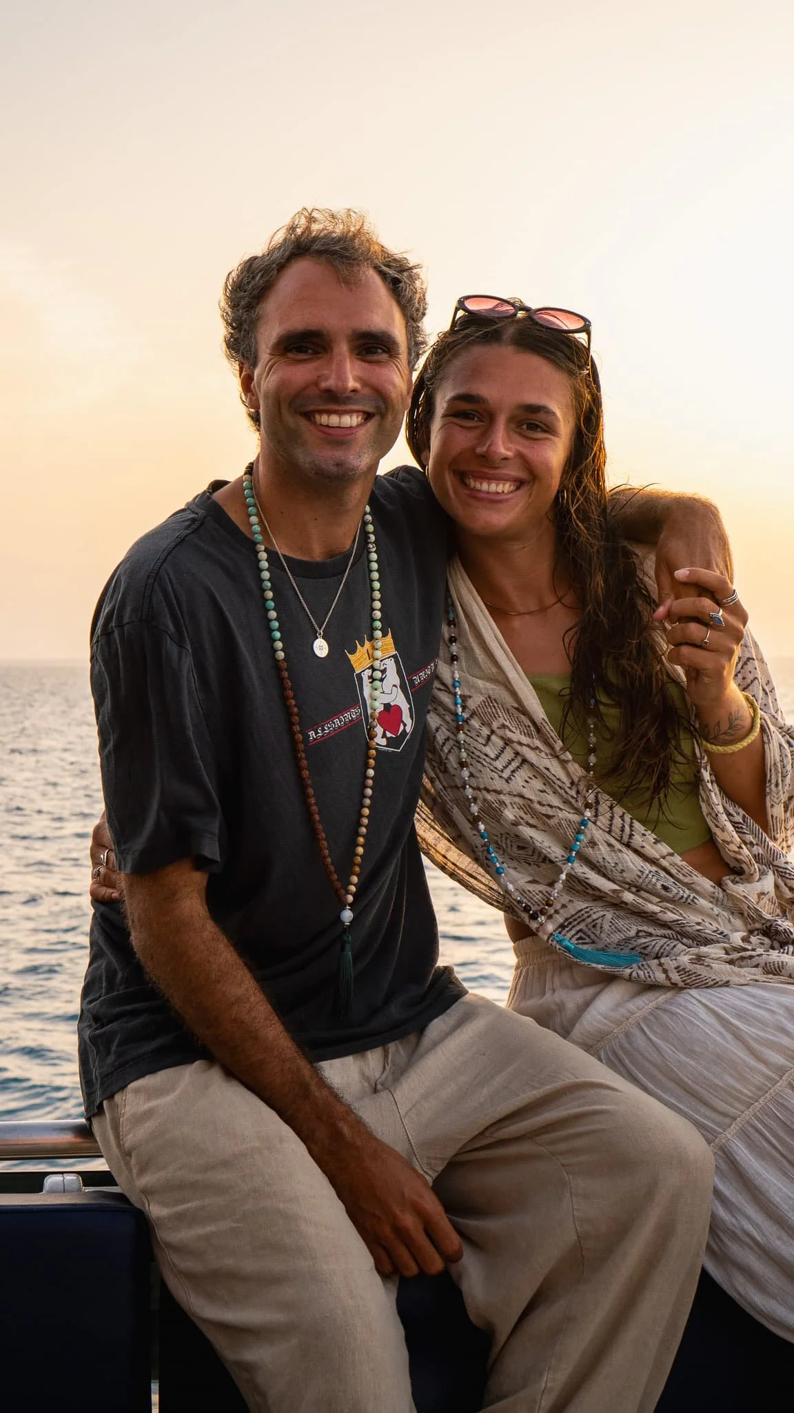 A smiling man and woman sitting on a boat at sunset, wearing casual clothing and beaded necklaces with the ocean in the background.