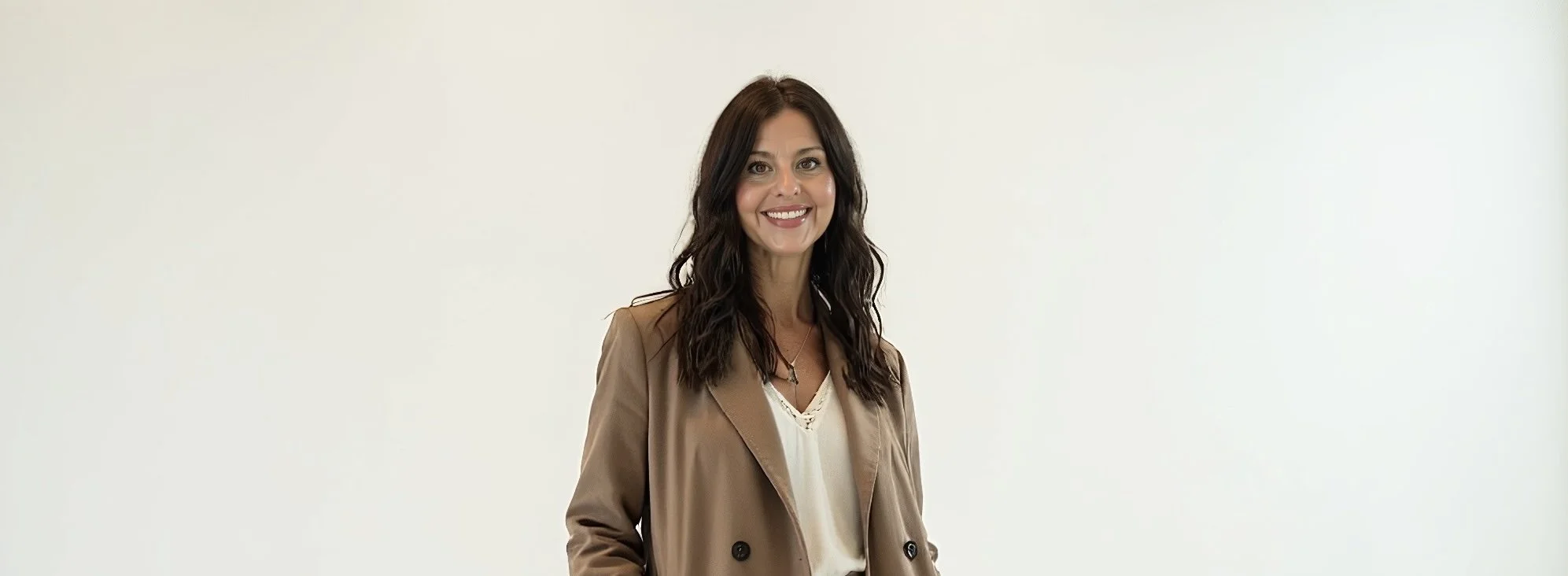 A woman with dark brown wavy hair, wearing a beige blazer over a white blouse, standing against a plain light background, smiling at the camera.