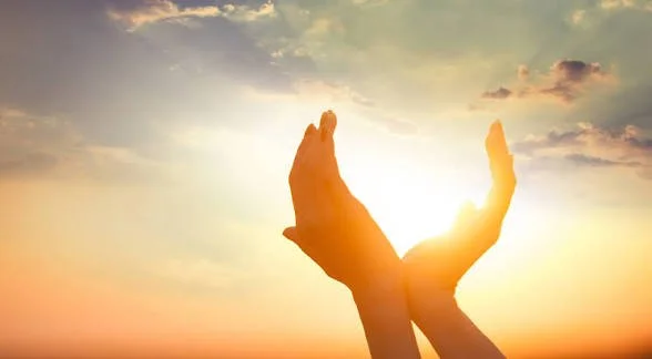 Two hands raised upward against a sunset sky, with the sun between the hands.