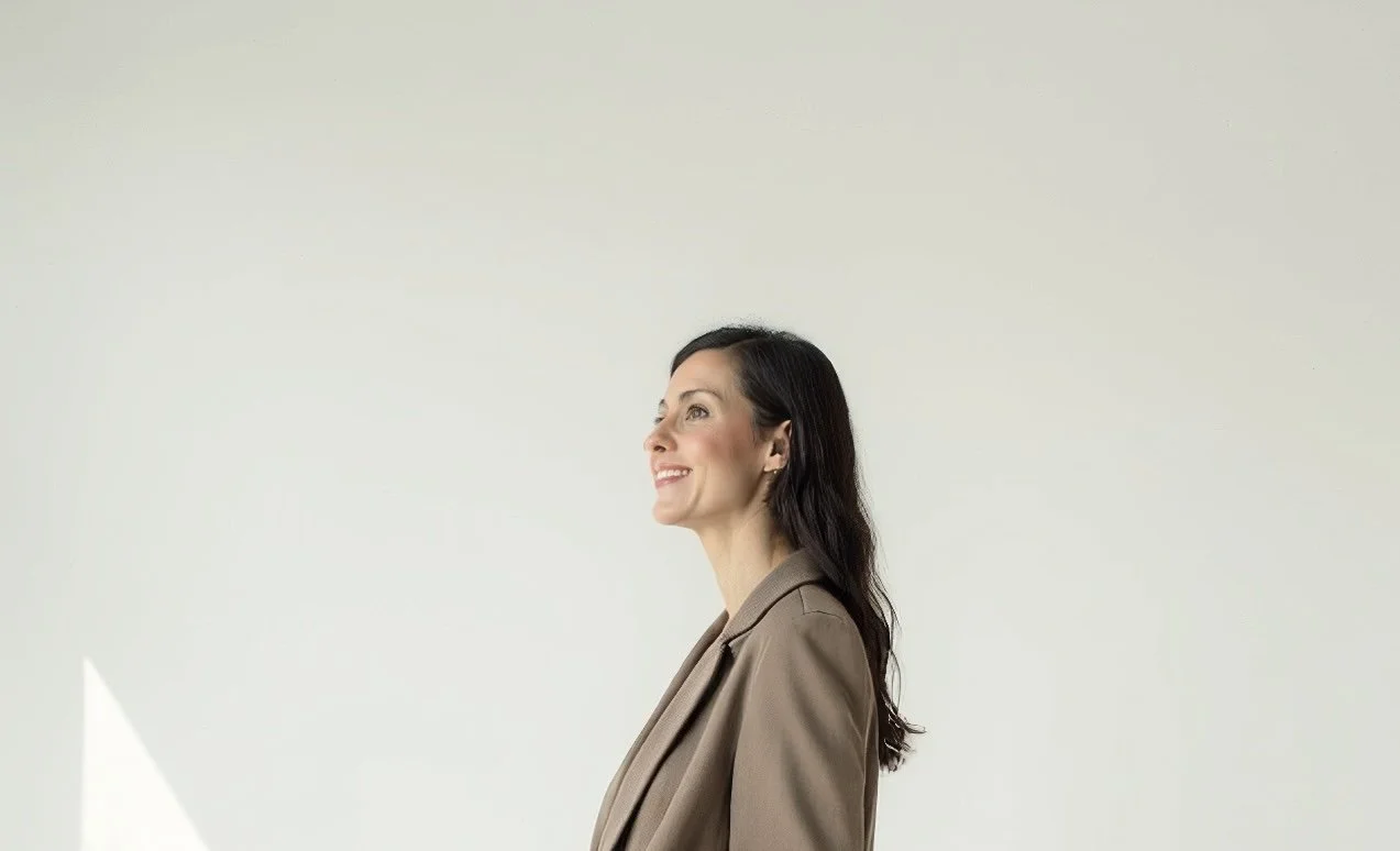 A woman with dark hair smiling and looking to her left, dressed in a beige blazer, standing against a plain light-colored background.