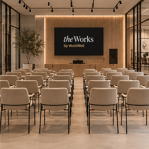 Conference room with beige chairs, large screen displaying 'theWorks by WorkWell,' potted plants, and glass walls.