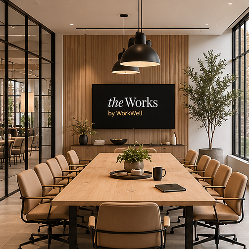 Conference room with a large wooden table, beige chairs, and a black sign that says 'the Works by WorkWell' on a wood-paneled wall. There are hanging pendant lights and potted plants, with large windows letting in natural light.