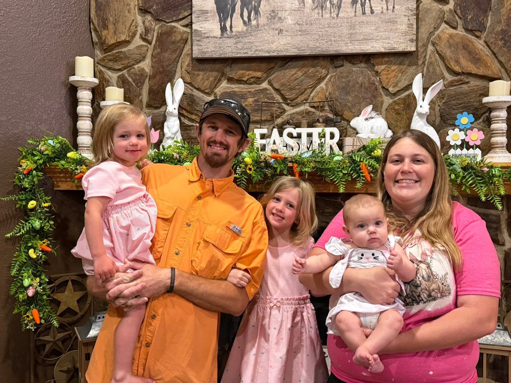 A group of six people, including three children, poses indoors in front of a stone fireplace decorated with Easter-themed ornaments and greenery. The group includes a man in an orange shirt, a woman in a pink shirt, and children with light hair and fair skin. There are bunny figurines and colorful signs that say 'Easter' and 'Hoppy Spring' in the background.