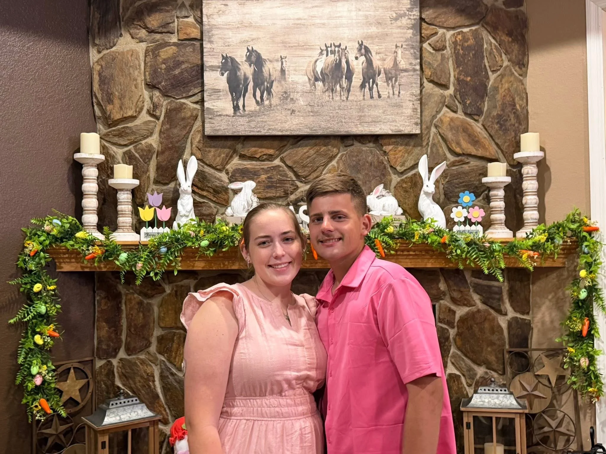 A young couple is standing in front of a decorated fireplace with a stone wall. The woman is wearing a pink dress, and the man is dressed in a pink shirt. The mantle is decorated with greenery, bunny figurines, and colorful paper flowers. Above the fireplace, there is a painting of horses walking on a plain.