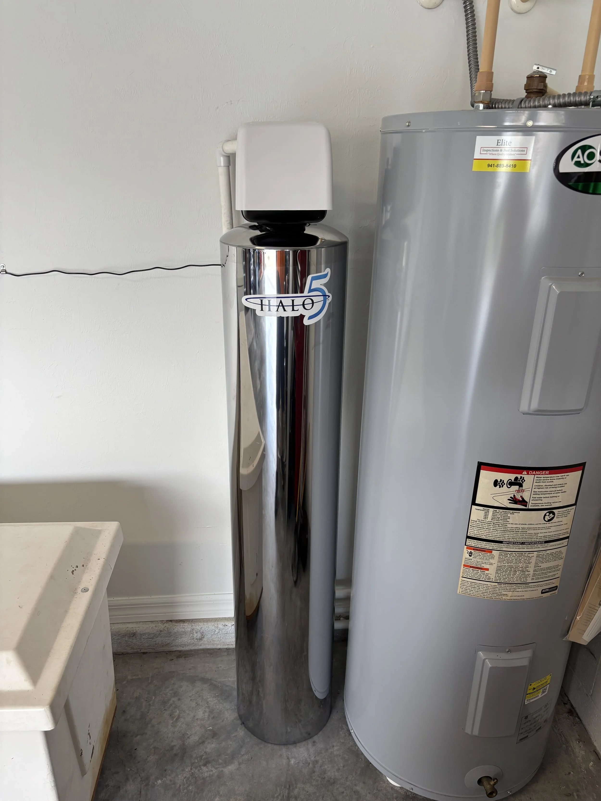 A household utility room with a water heater on the right, a polished metal water softener tank labeled 'HALO 5' in the middle, and a small white appliance on the left. The room has a gray concrete floor and white walls.