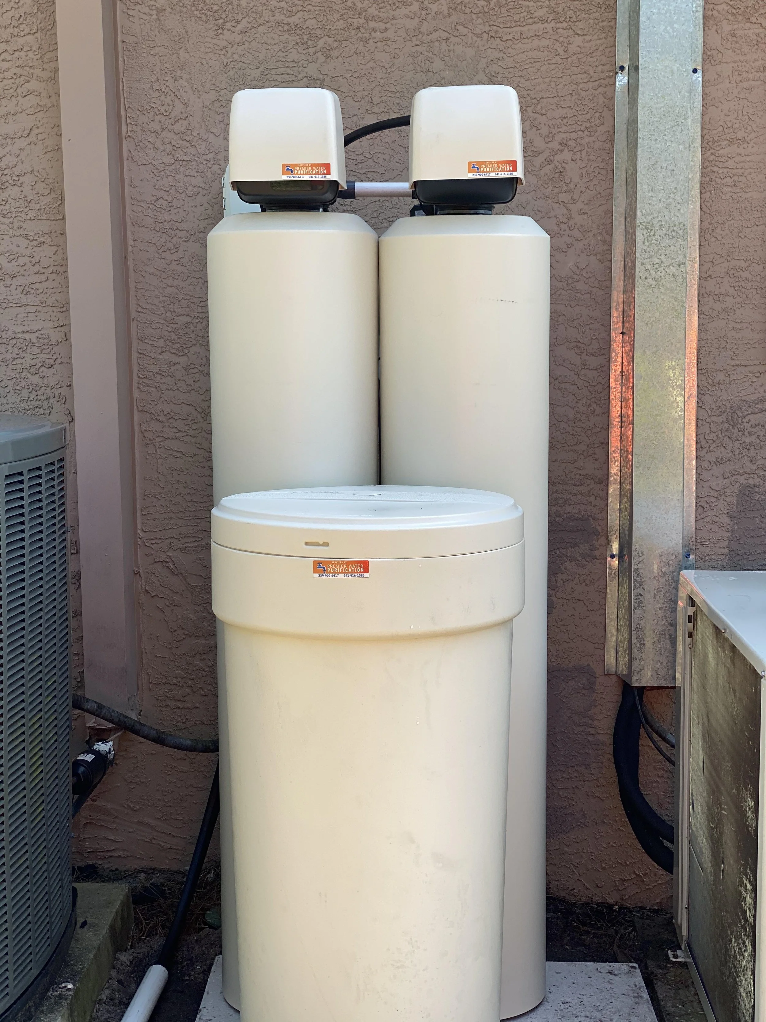 Two large water filtration units are positioned outside against a textured beige wall, with stickers indicating premier water purification installed and maintain the equipment. 