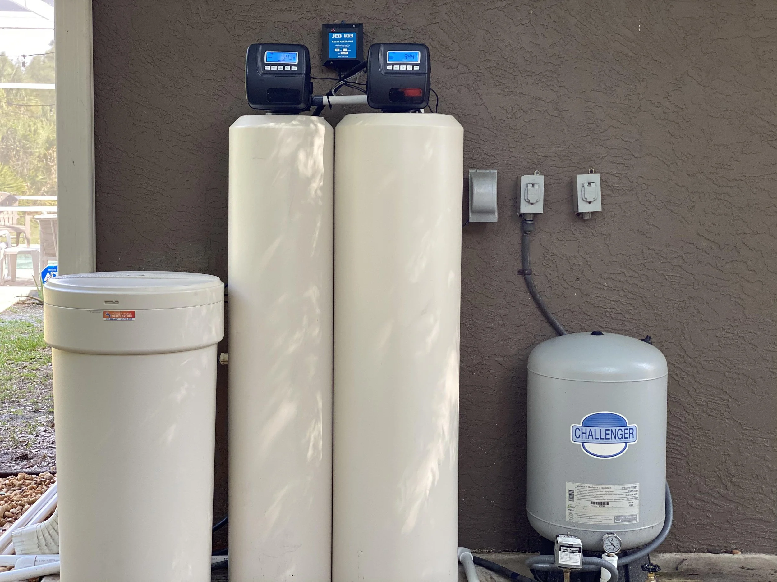Water filtration system with white cylindrical filters, digital control panels, and a pressure gauge mounted on a beige stucco wall outdoors.