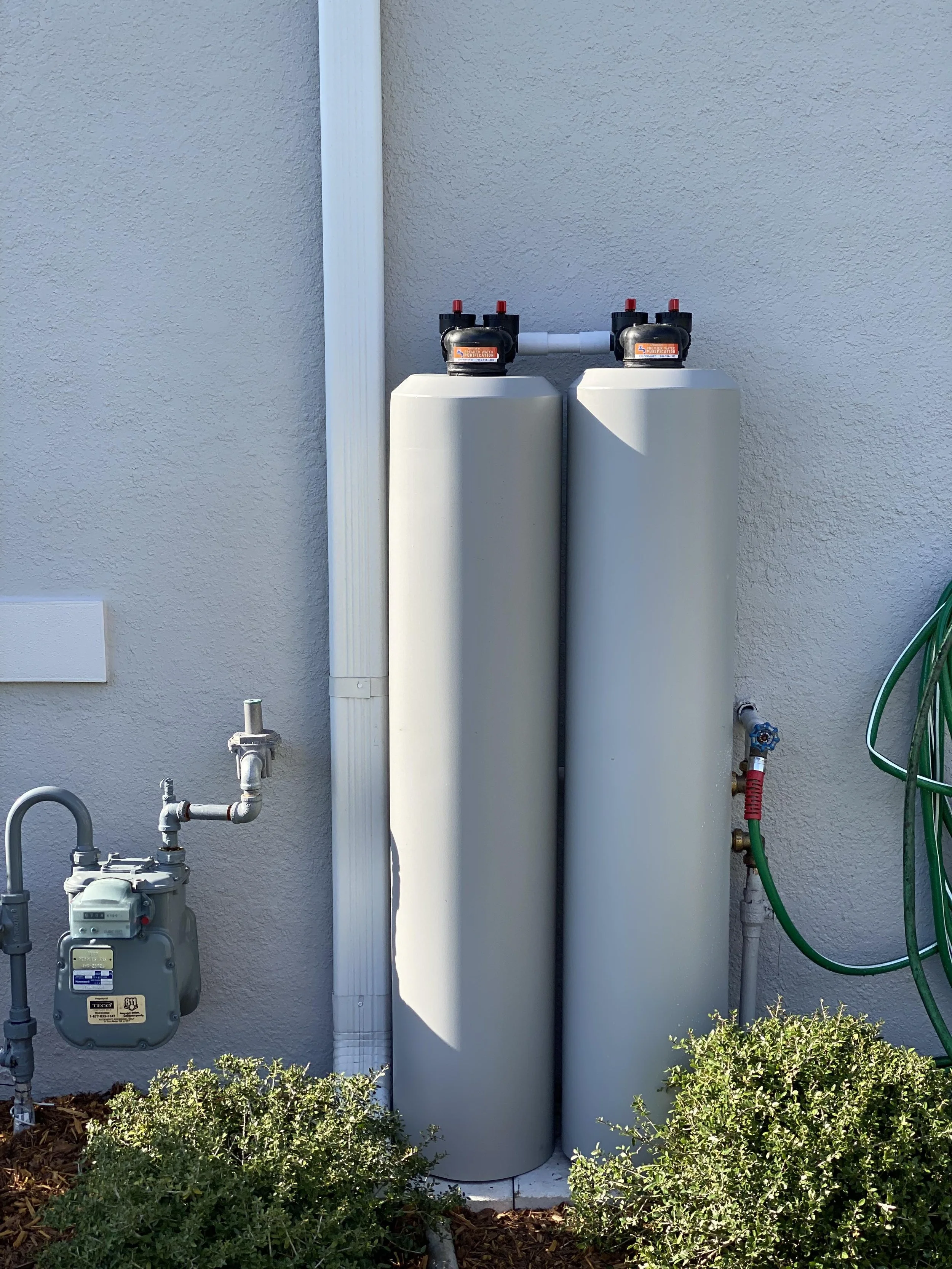 Outside view of a residential wall with two water filter tanks, pipes, with small bushes at the bottom.